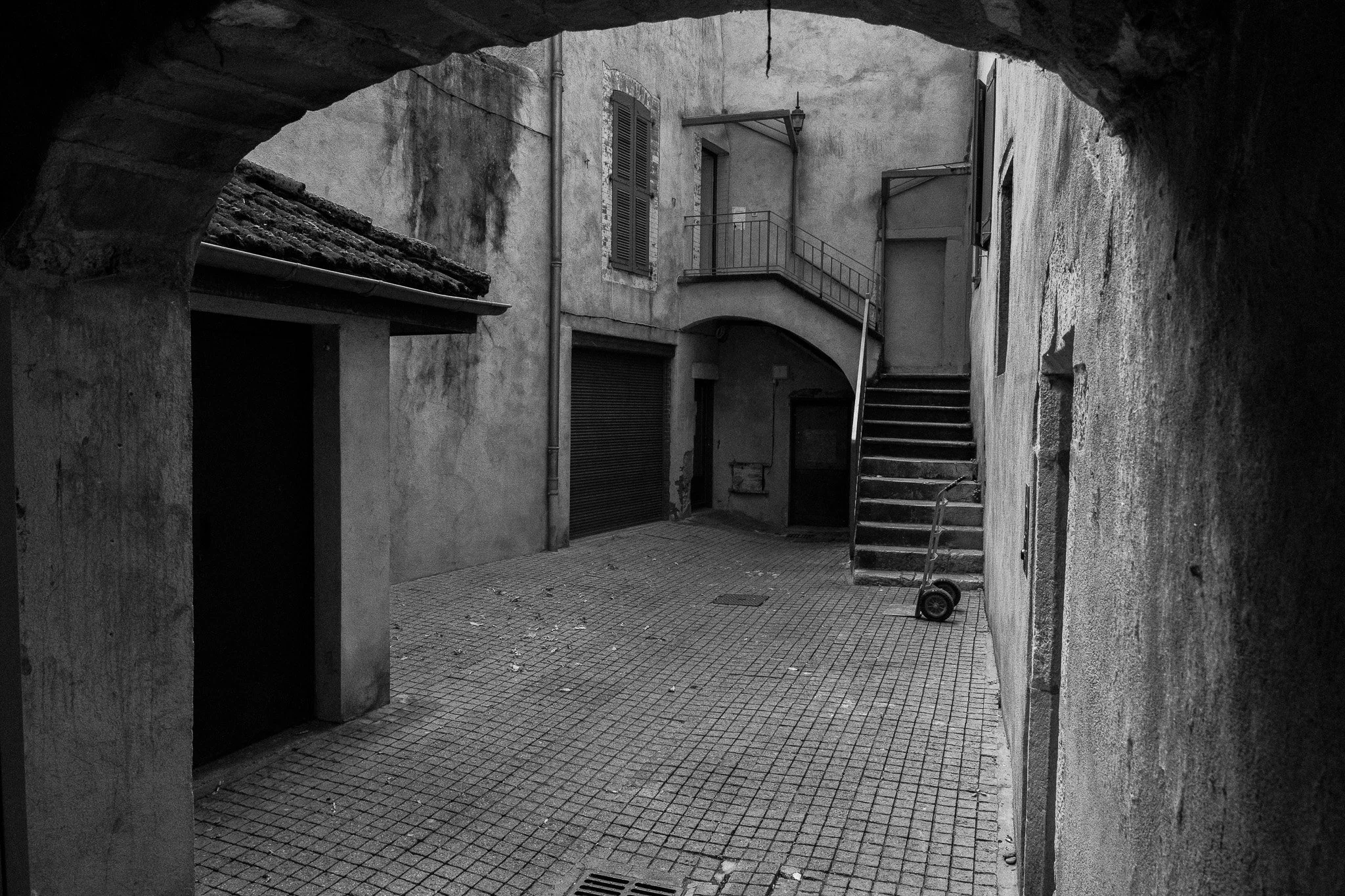Une cour intérieure d'immeuble ancien en noir et blanc, avec des escaliers en spirale, des fenêtres à volets, une pompe à main et un scooter électrique.