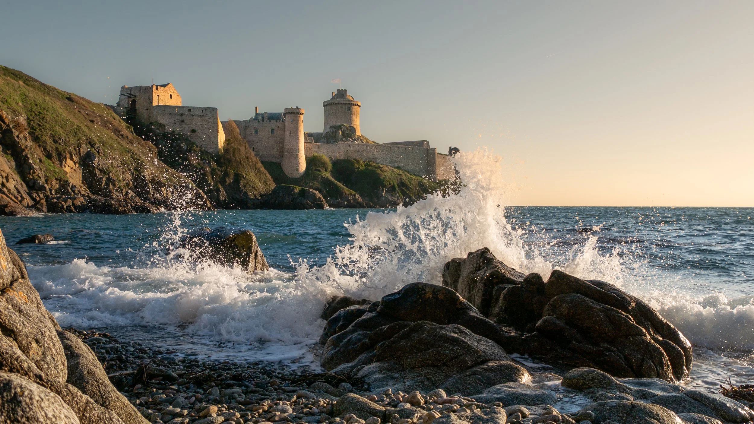 Château fortifié perché sur une falaise rocheuse au bord de la mer, avec des vagues frappant les rochers en premier plan, en fin d'après-midi
