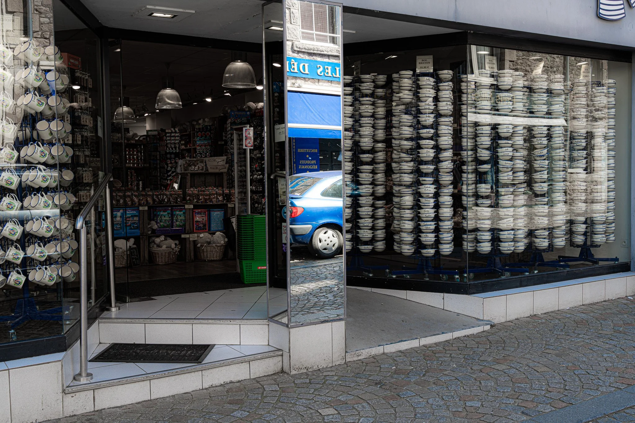 Vitrine d'une boutique de souvenirs avec des mugs et des assiettes en céramique, certains avec des inscriptions en français, et une voiture bleue garée dehors. La vitrine est en verre et la boutique se trouve dans une rue pavée.