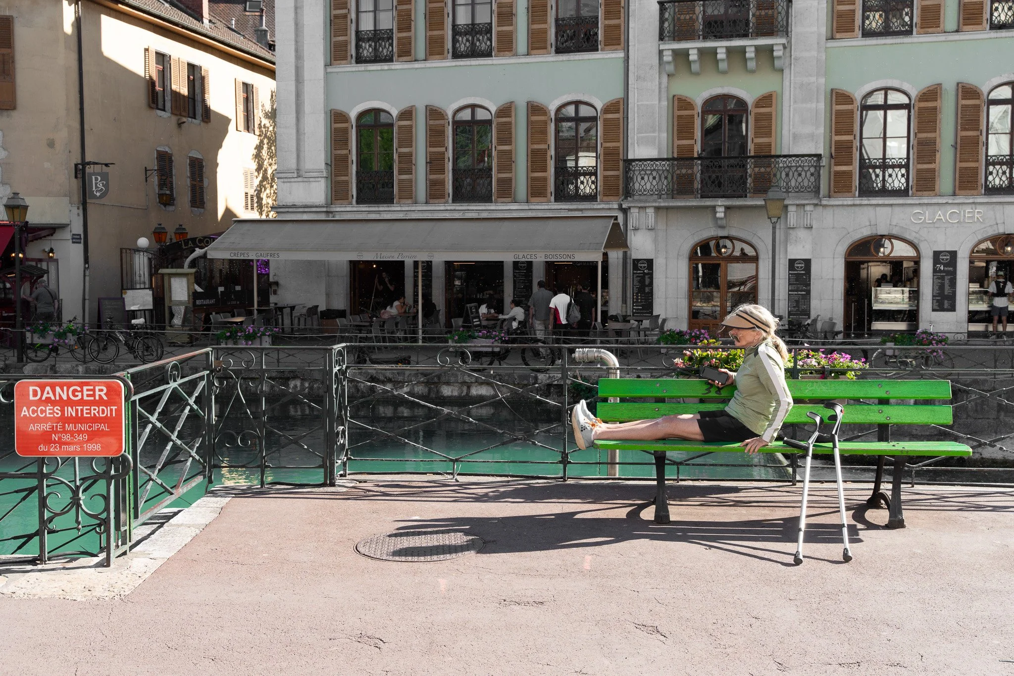 Une femme assise sur un banc vert au bord d'un canal, portant un chapeau et une veste claire. En arrière-plan, il y a plusieurs bâtiments avec des fenêtres à volets en bois et une terrasse où des personnes mangent. Il y a aussi une enseigne pour une crêperie et un panneau rouge avec une interdiction d'accès.