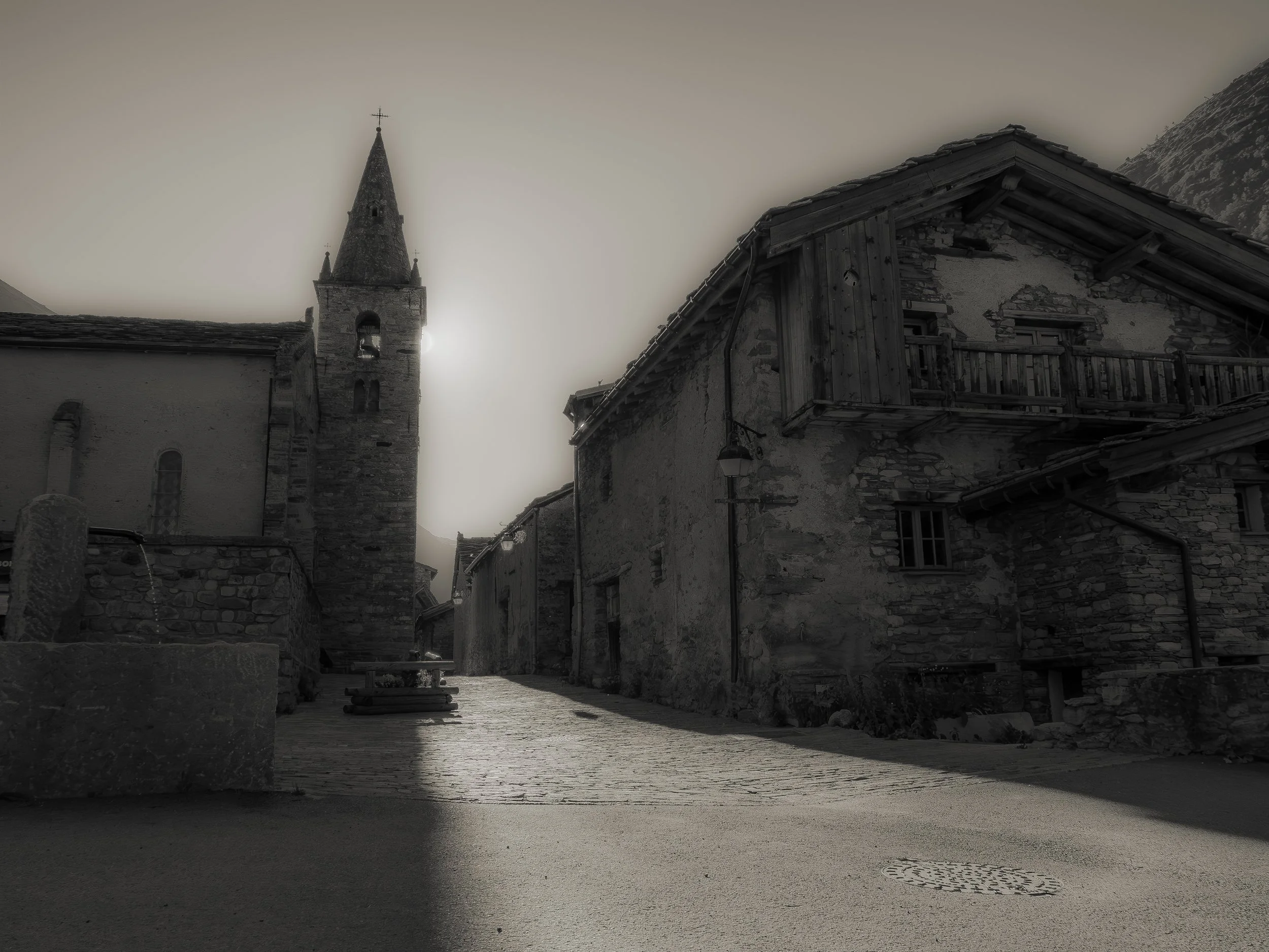 Rue pavée dans un village de montagne, avec des bâtiments en pierre et un clocher d'église en arrière-plan, sous un ciel nuageux, en noir et blanc.