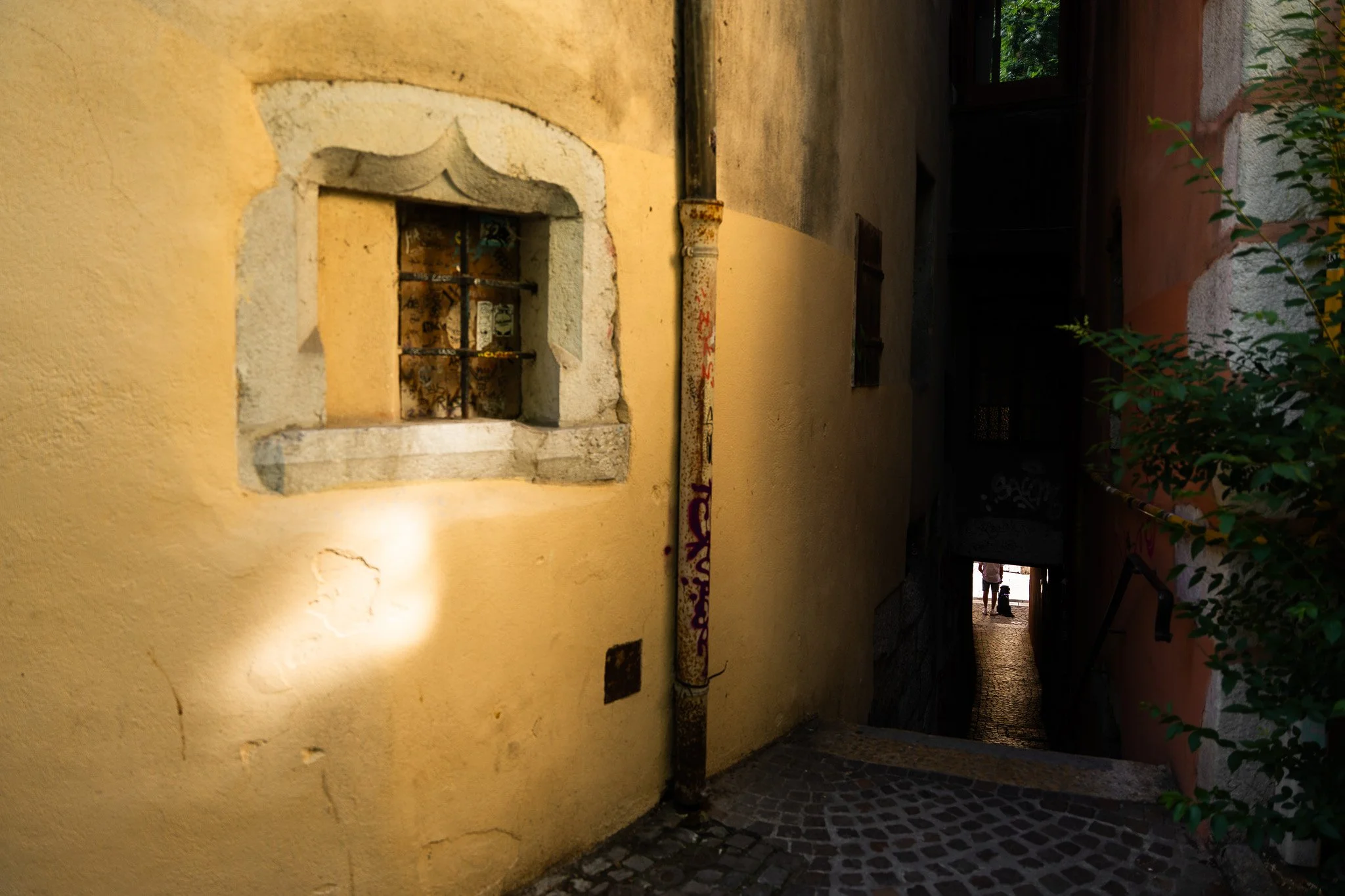Photo montrant un mur jaune avec une petite fenêtre en pierre avec barreaux métalliques, un tuyau rouillé traversant la scène, et une entrée ouverte au fond. Il y a des plantes à droite et deux personnes visibles dans le fond, sous un ciel clair.