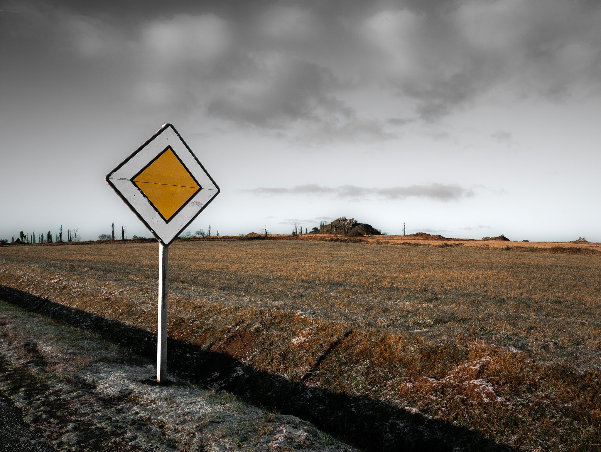 Un panneau de signalisation routière jaune avec un symbol de virage, placé dans un paysage rural avec un ciel nuageux et des champs ouverts.