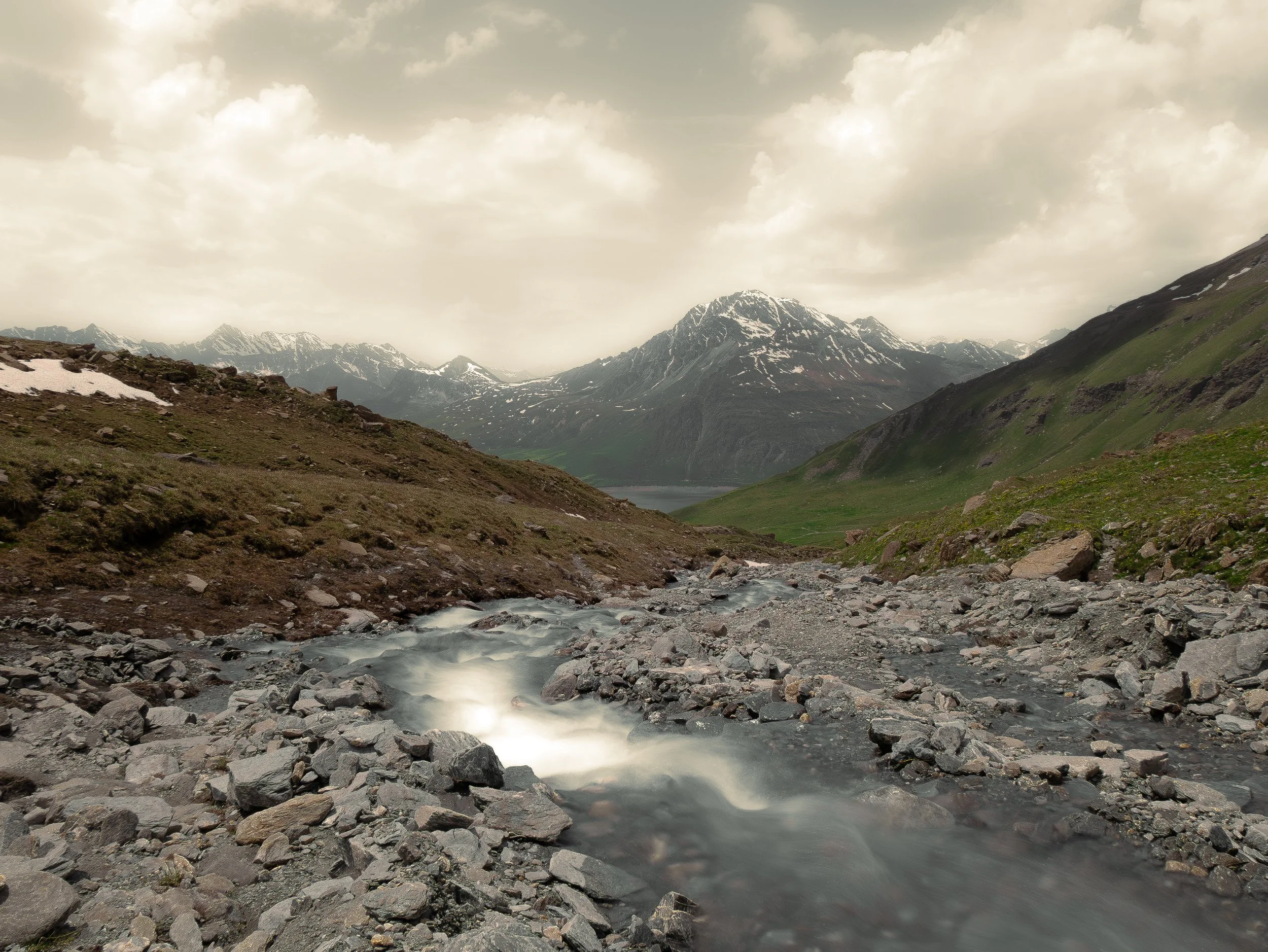 Montagnes enneigées vue d'une vallée avec un ruisseau rocheux en premier plan et un ciel nuageux.