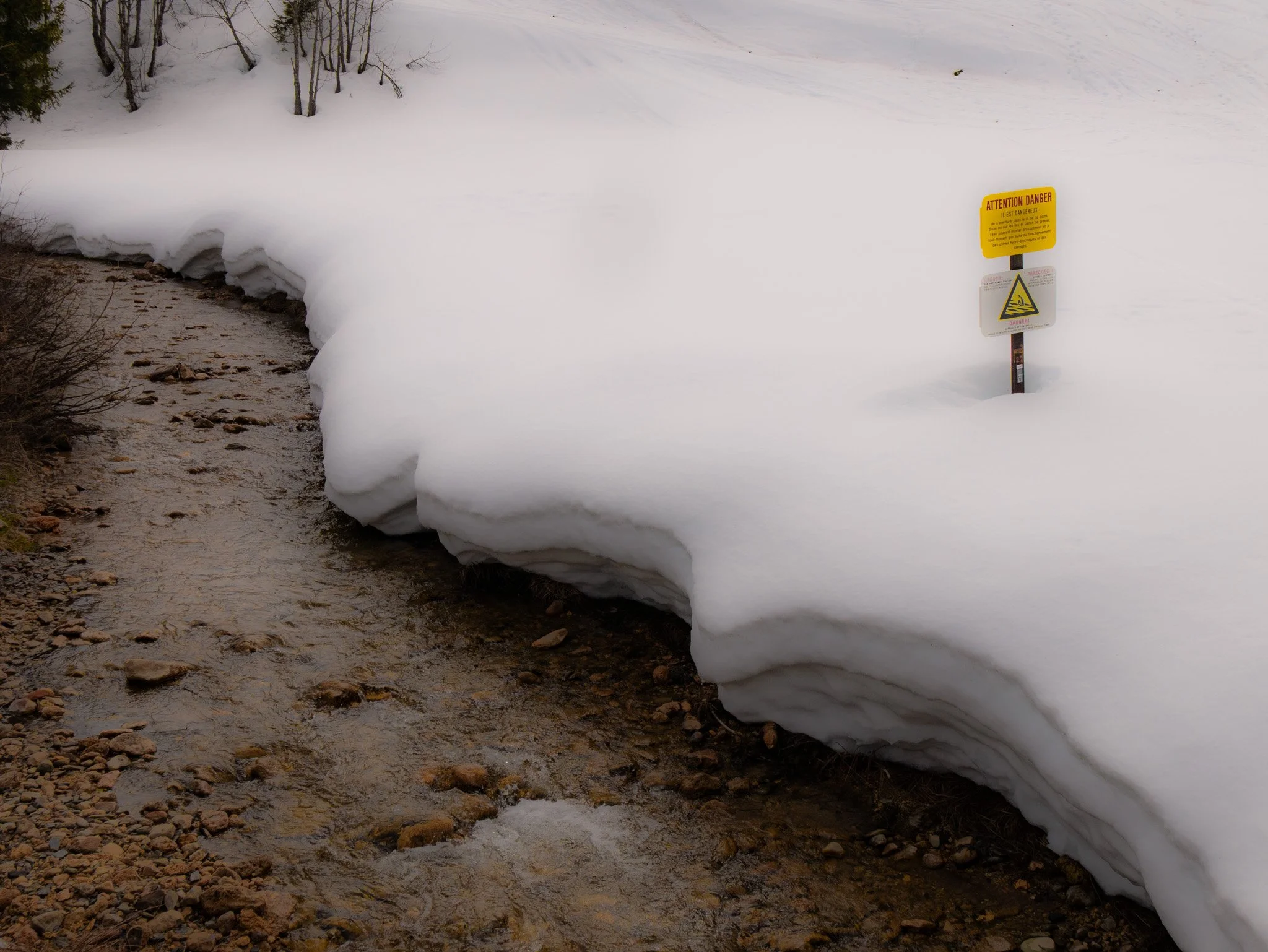 Une rivière coule sous une couche épaisse de neige en hiver. Un panneau d'avertissement en français indique un danger potentiel dû à la glace ou à la chute dans l'eau.