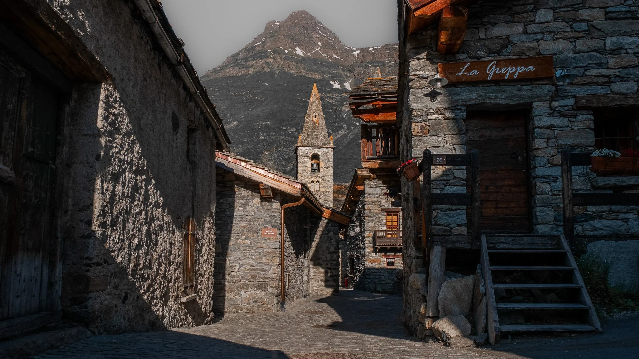 Rue pavée dans un village de montagne avec des maisons en pierre, un clocher d'église, et une montagne escarpée en arrière-plan, sous un ciel brumeux.