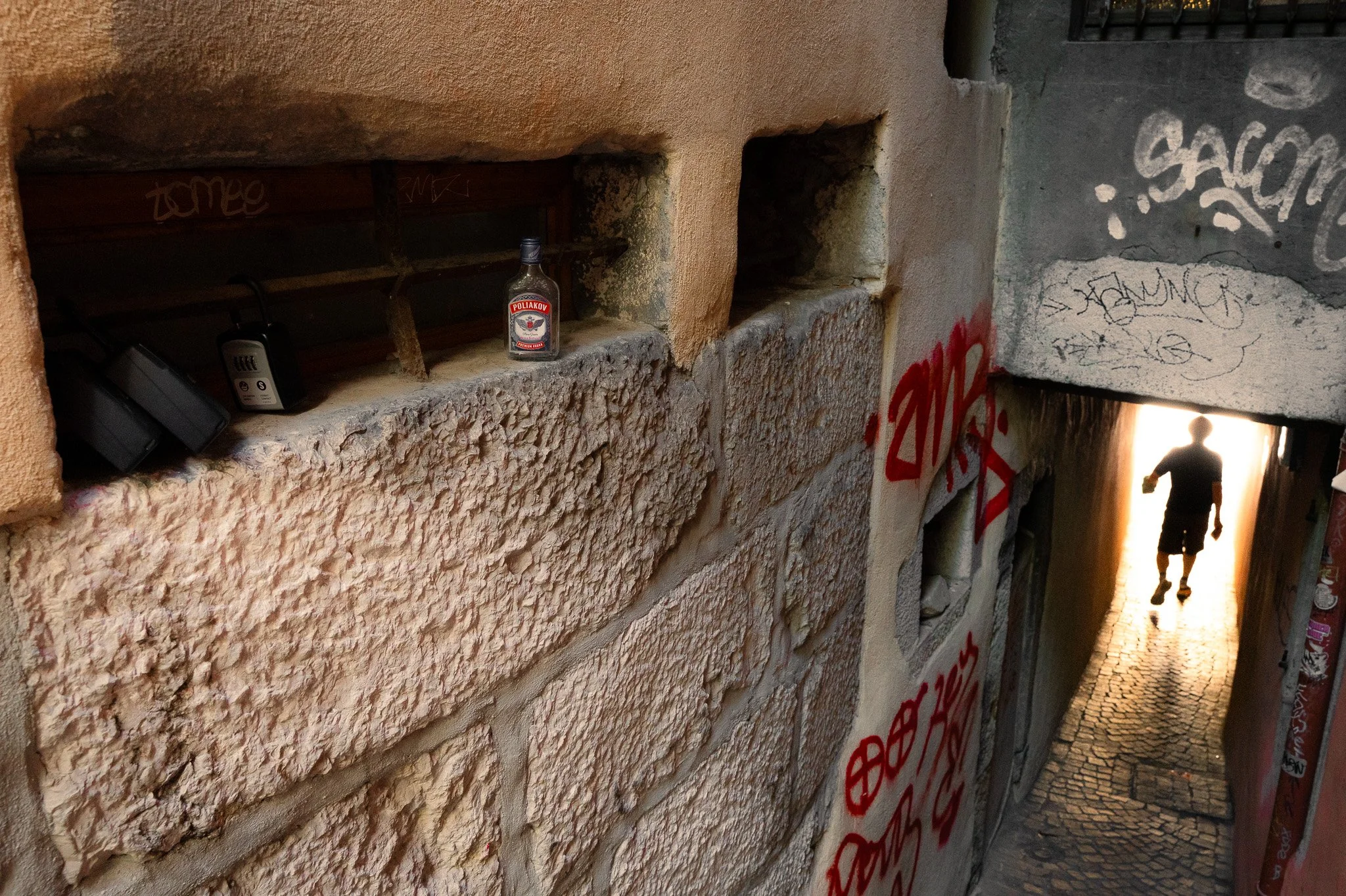 Un mur en pierre avec une petite fenêtre contenant une bouteille et deux appareils électroniques, dans une ruelle pavée avec un passage sous un pont.