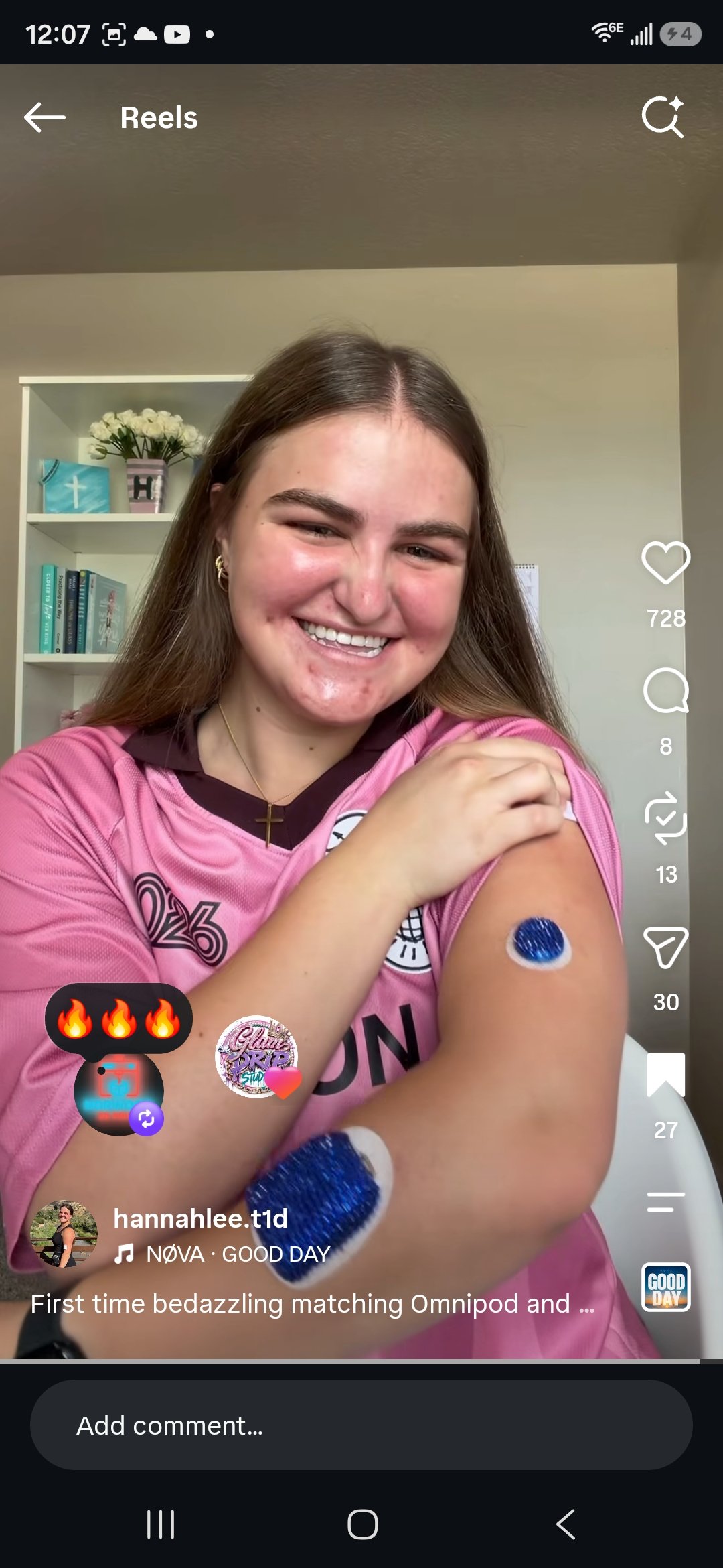 Young woman smiling and showing off her arm with a blue glittery tattoo, wearing a pink sports jersey, inside a room with a white bookshelf and flowers in the background