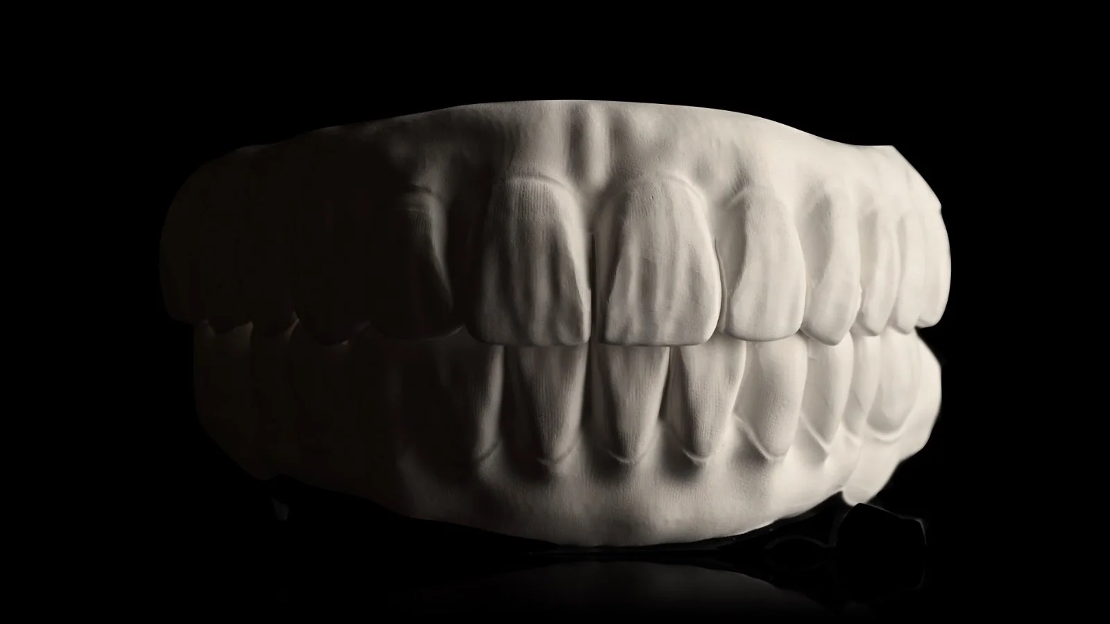A realistic sculpture of human teeth with both upper and lower rows, lit from above, against a black background.