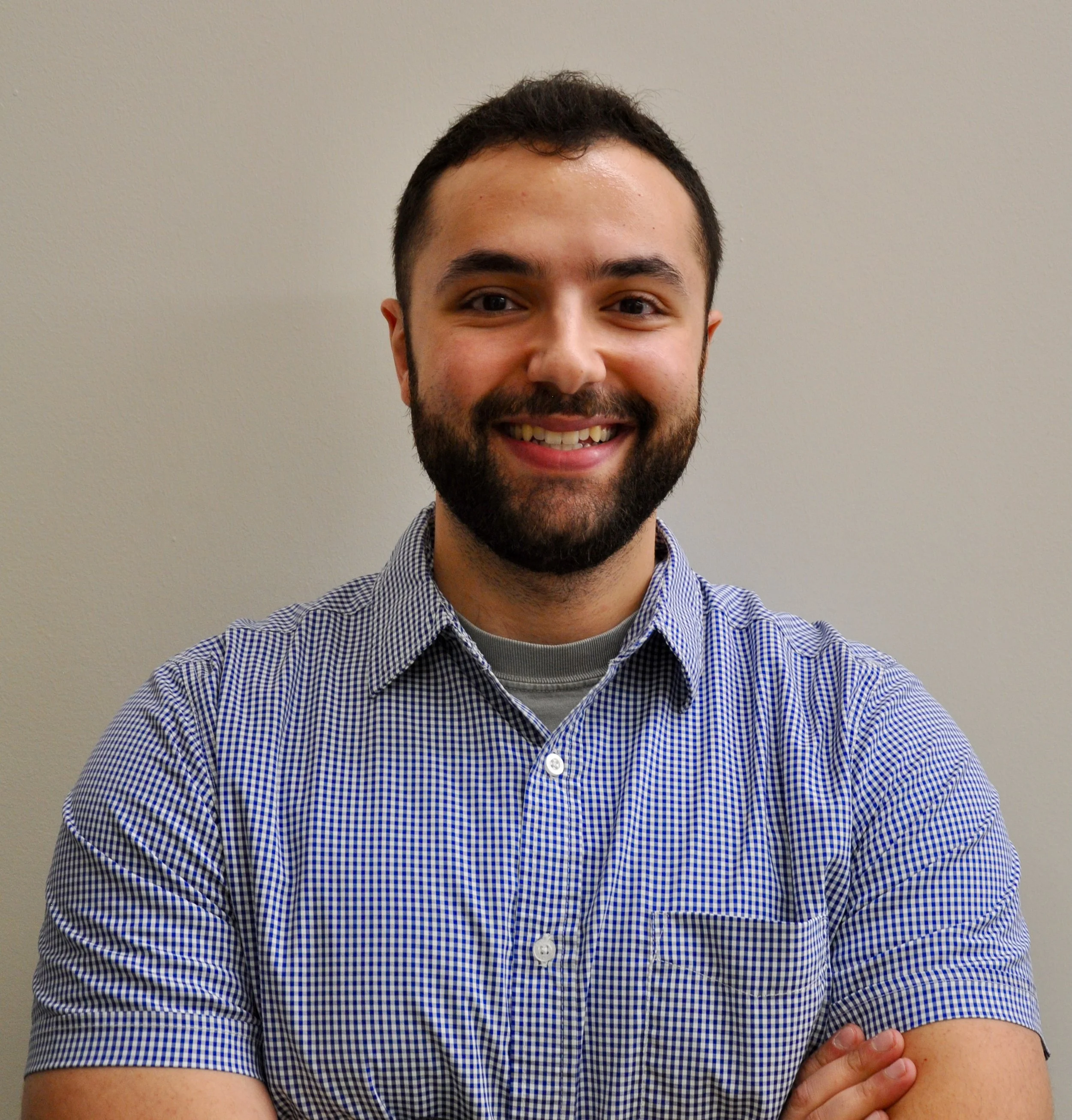 A young man with a beard and dark hair smiling, wearing a blue and white checkered shirt, standing against a plain light-colored wall.