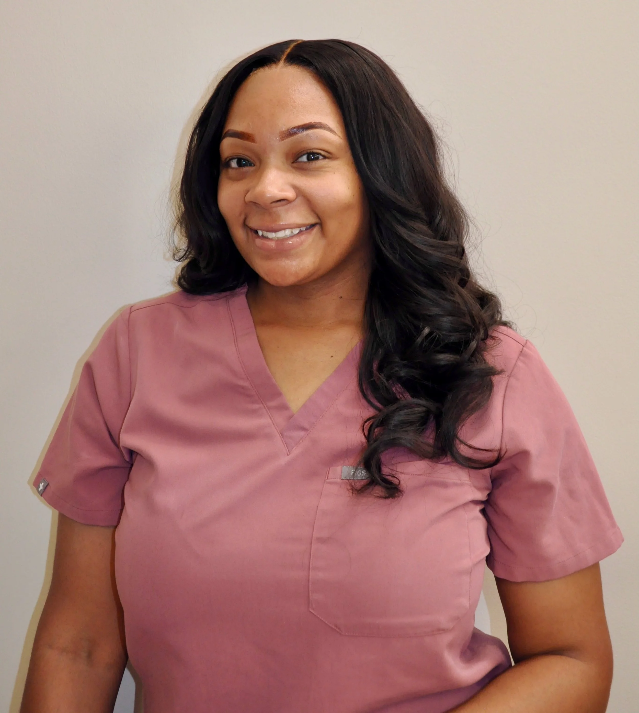 A woman in pink scrubs smiling, with long, dark wavy hair and a light background.