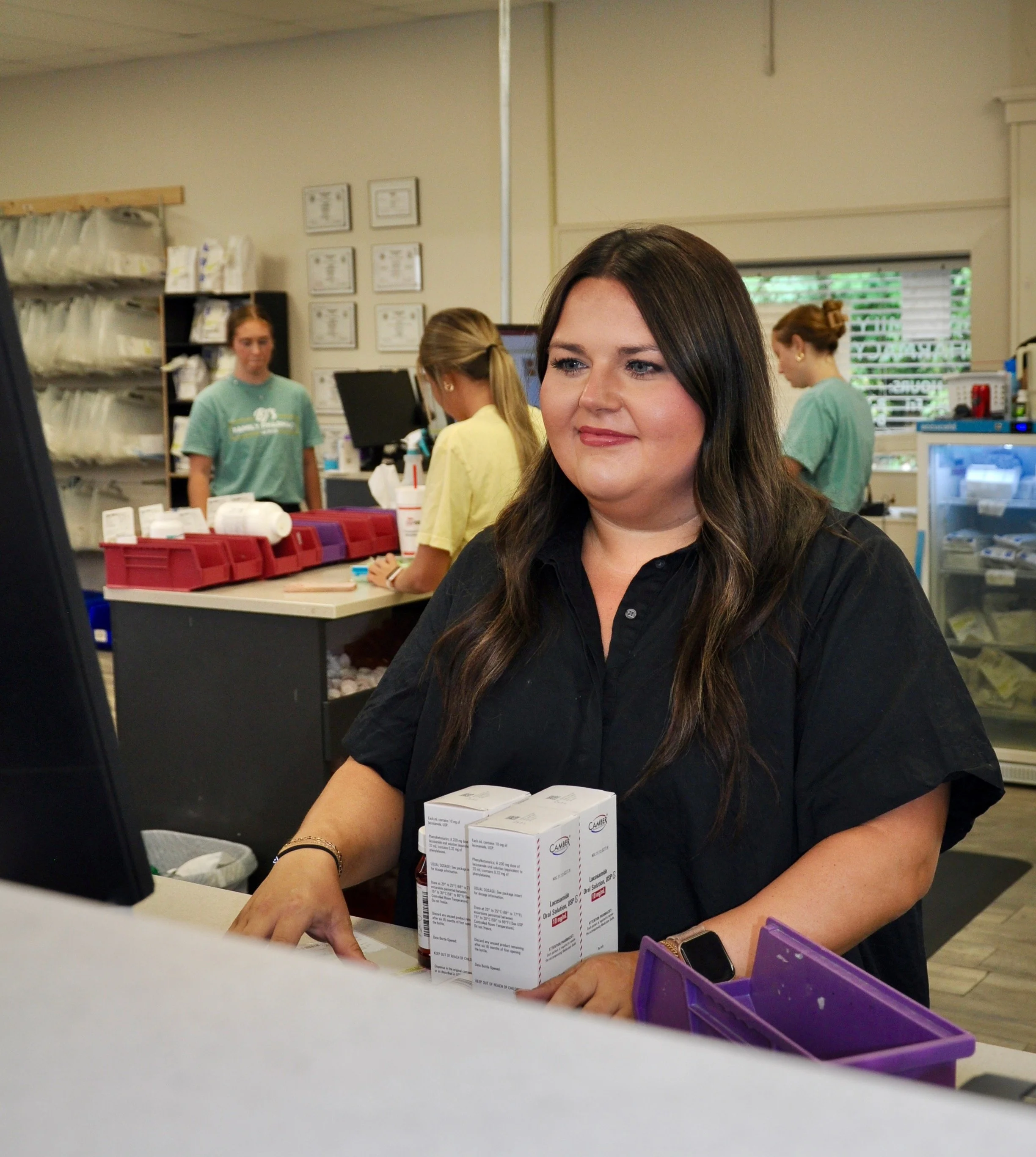 A woman with long dark hair working at a pharmacy counter, with pharmacy staff in turquoise and yellow scrubs in the background. Shelves with packaged supplies and certificates on the wall are visible inside a pharmacy.
