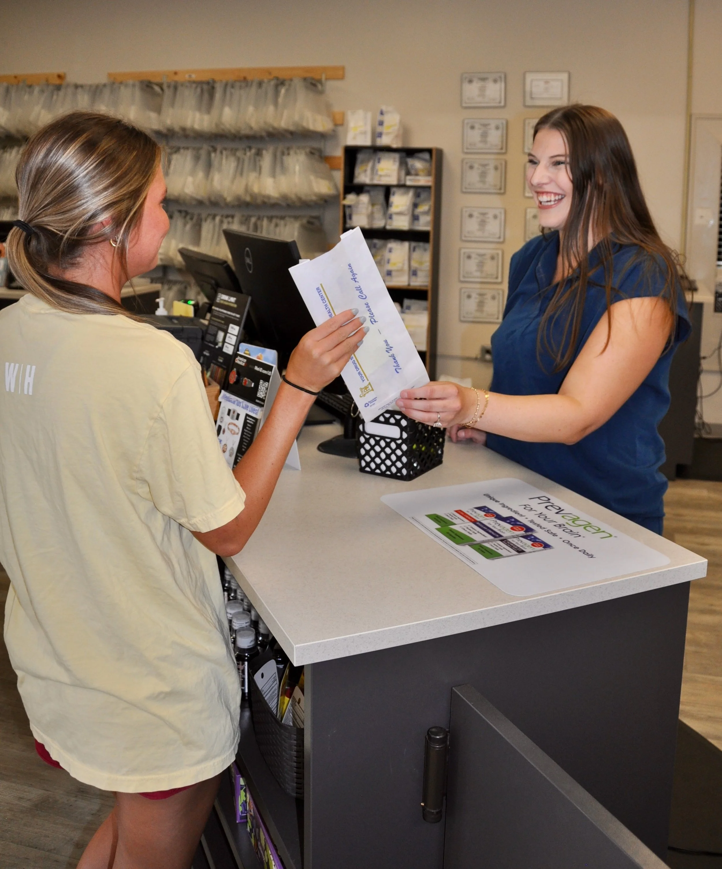 A young woman with long brown hair and a big smile at a counter, handing over an envelope to a cashier in a retail store. The cashier sports a beige t-shirt, and the background features shelves with various items and framed certificates on the wall.