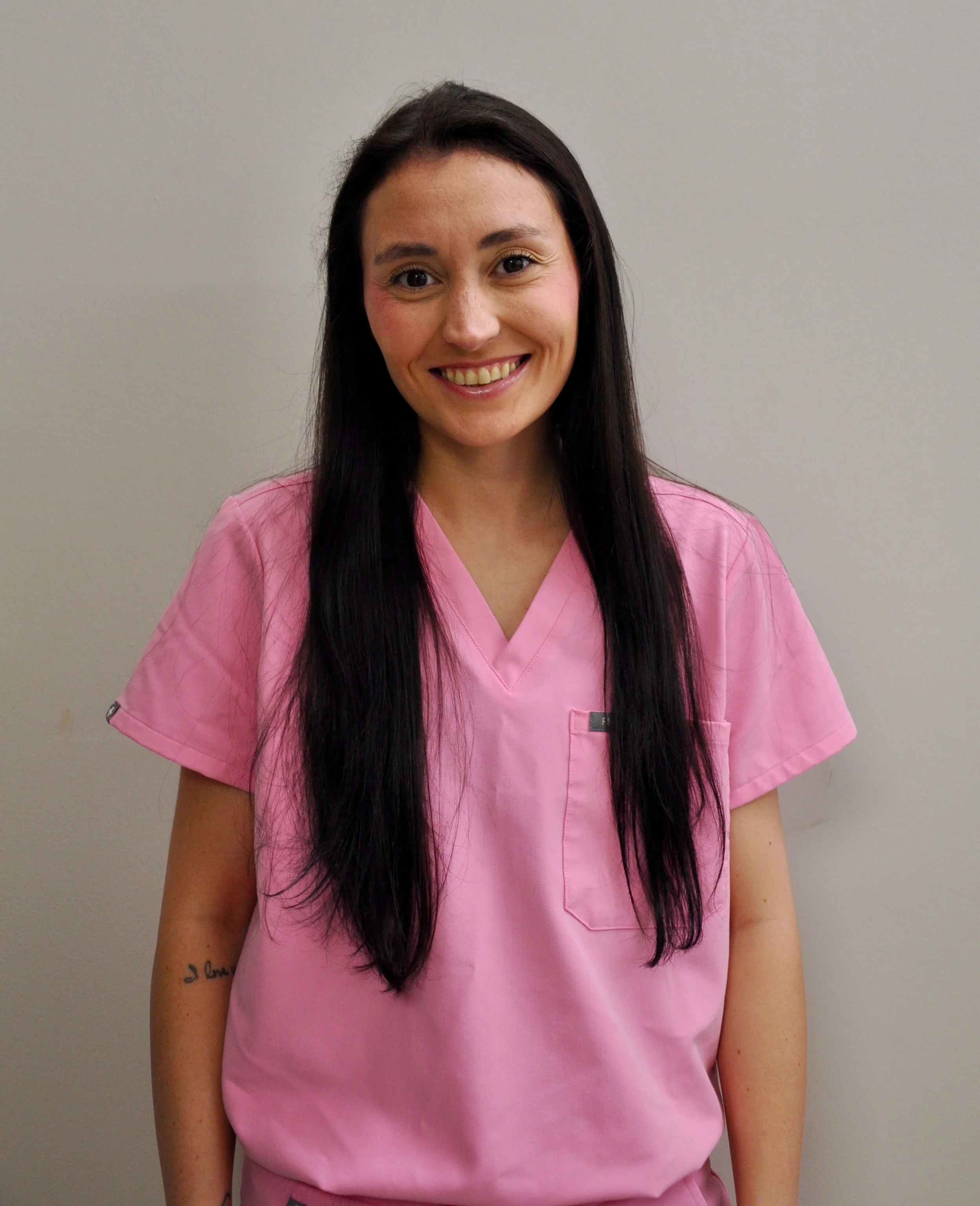 A woman with long dark hair wearing light pink medical scrubs, smiling, standing against a plain gray wall.