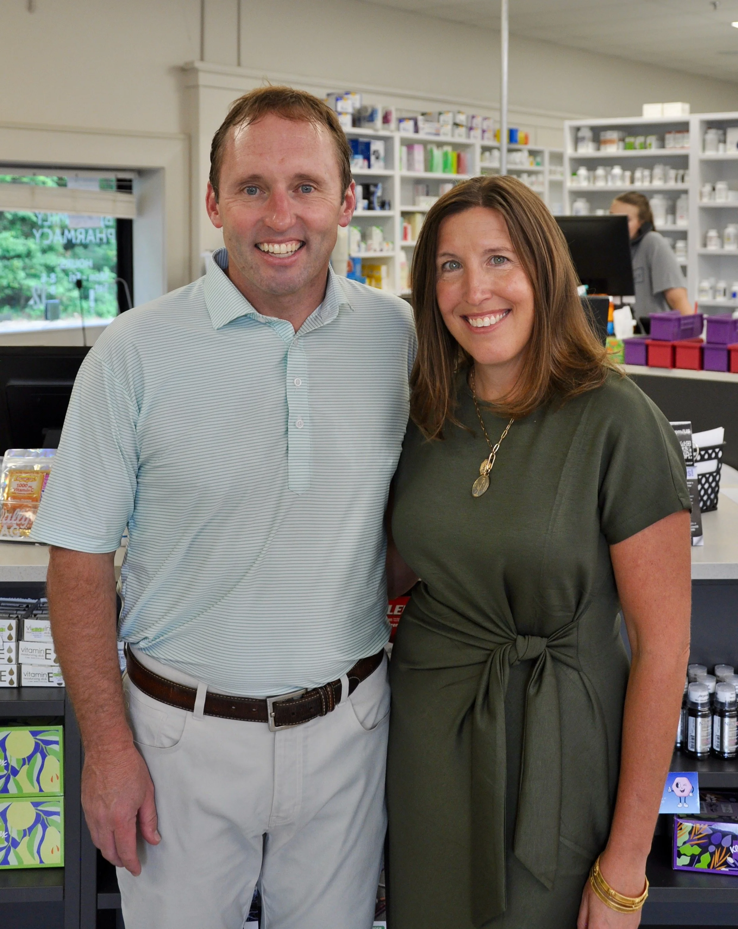 A man and woman standing close together in a pharmacy, smiling at the camera with pharmacy shelves and a cashier in the background.