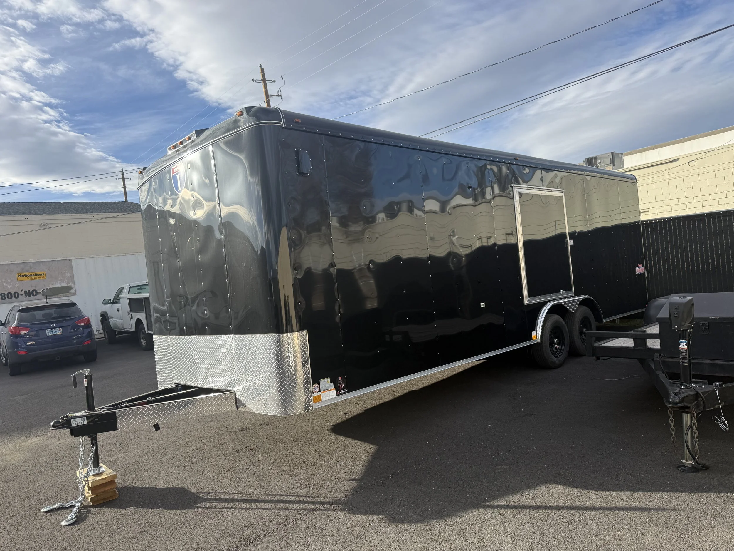 A shiny black enclosed trailer parked outdoors with a metal diamond plate front apron. The trailer is hitched to a tow vehicle and is next to other vehicles and a building, under a sky with scattered clouds.