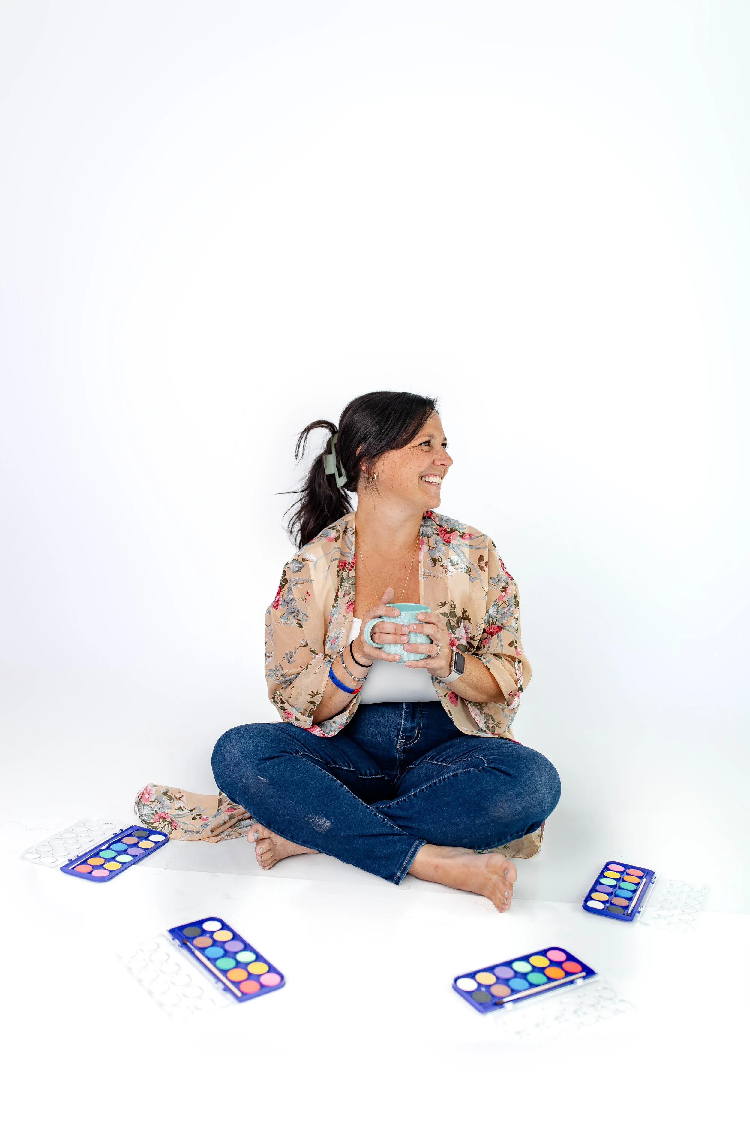 A woman sitting cross-legged on the floor against a white background, holding a mug and smiling, surrounded by open palettes of colorful makeup eyeshadows.