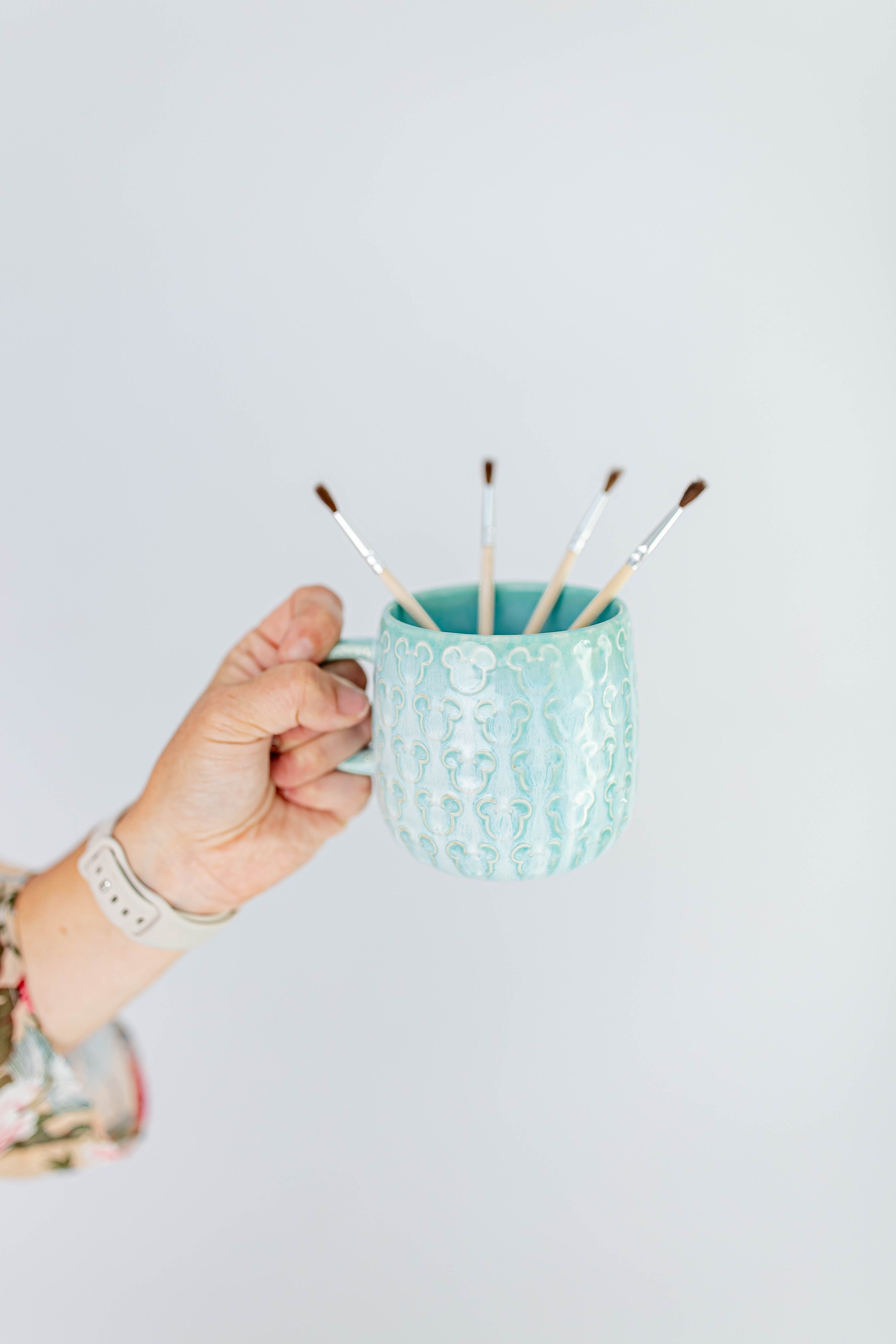 A person's hand holding a blue ceramic mug with a pattern and five paintbrushes inside it, against a plain light background.