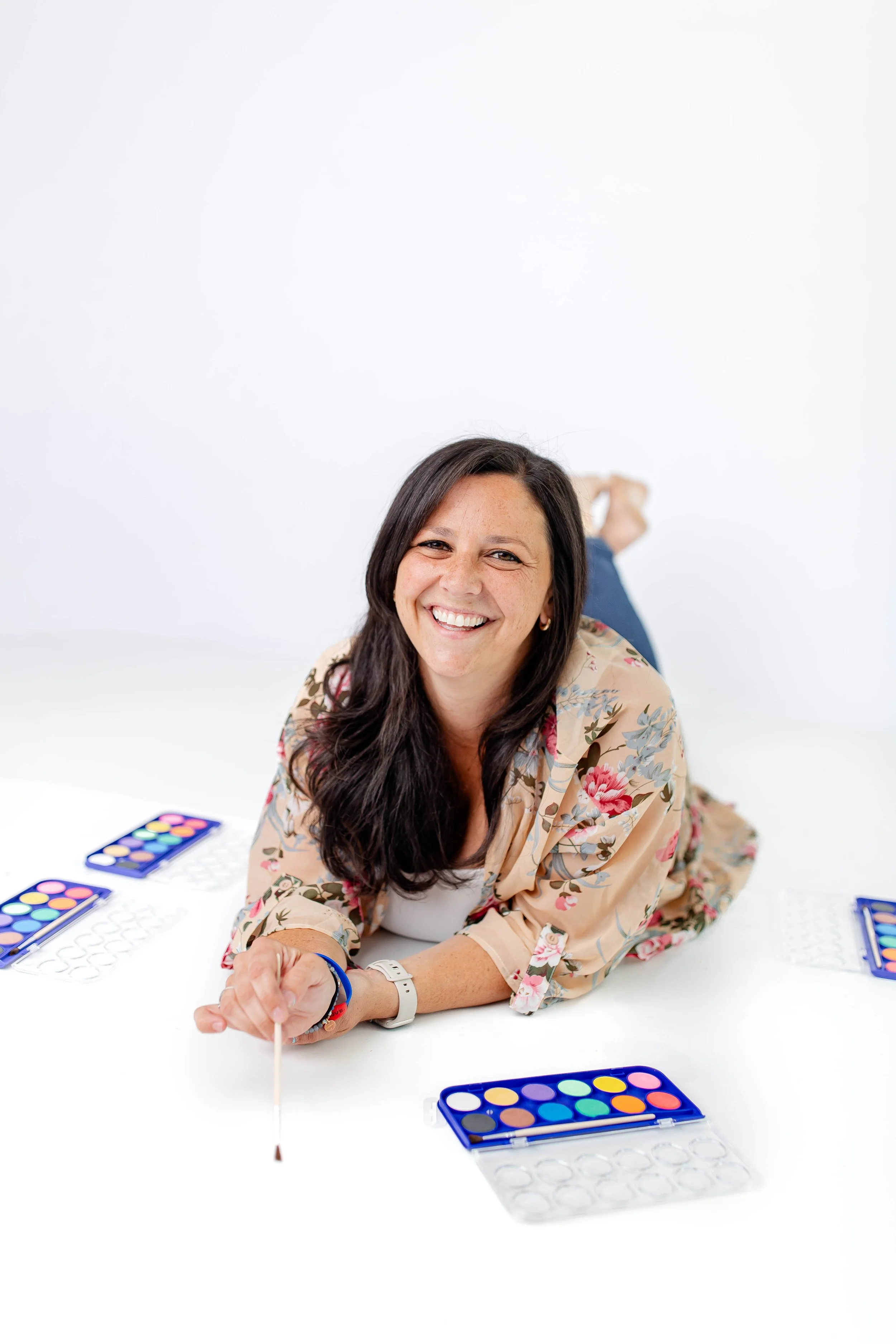 A woman with long dark hair and a floral shirt smiling while lying on a white table, holding a paintbrush with colored watercolor paints in front of her. It appears she is participating in a creative activity.