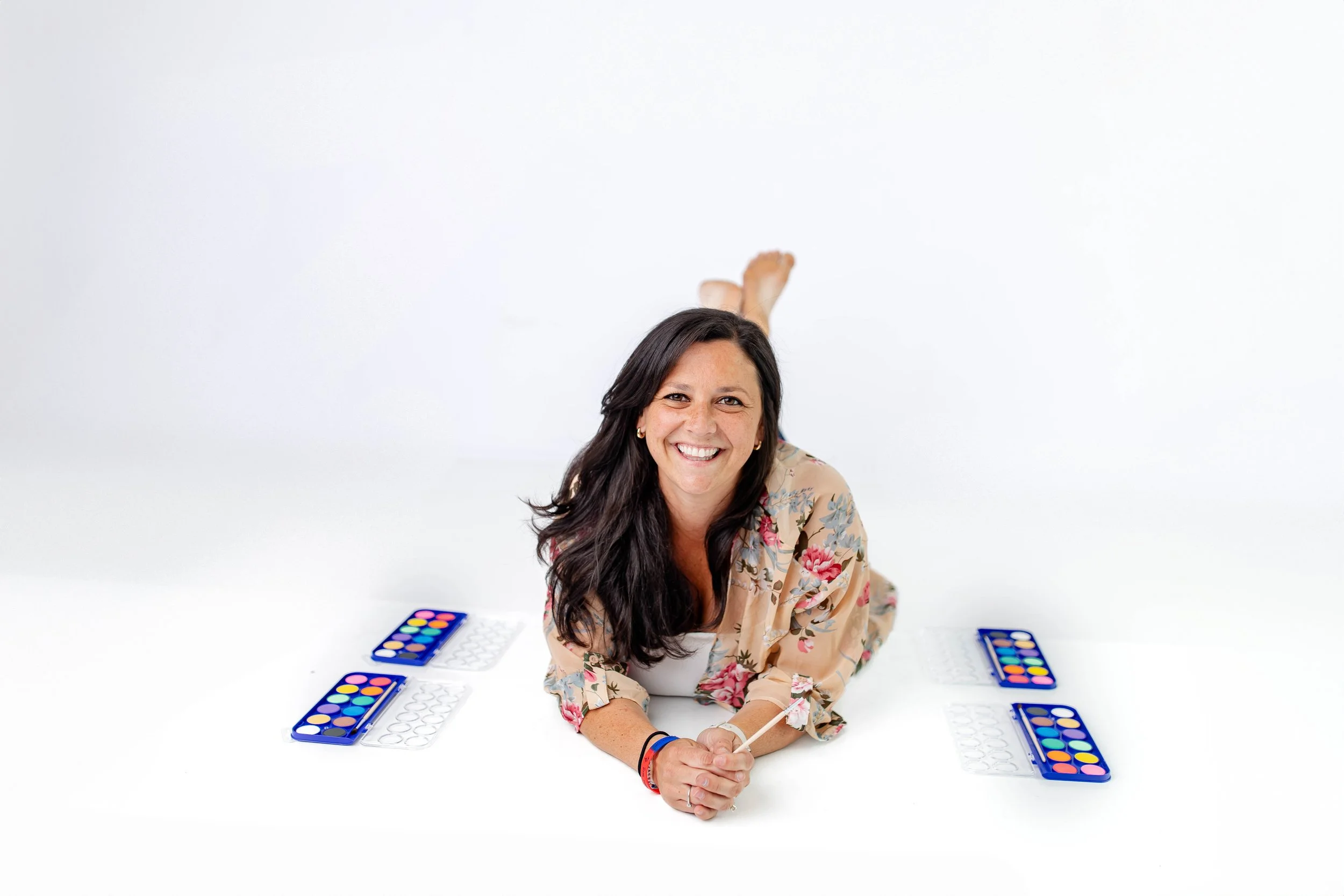 Woman smiling lying on a white surface surrounded by watercolor paint palettes.