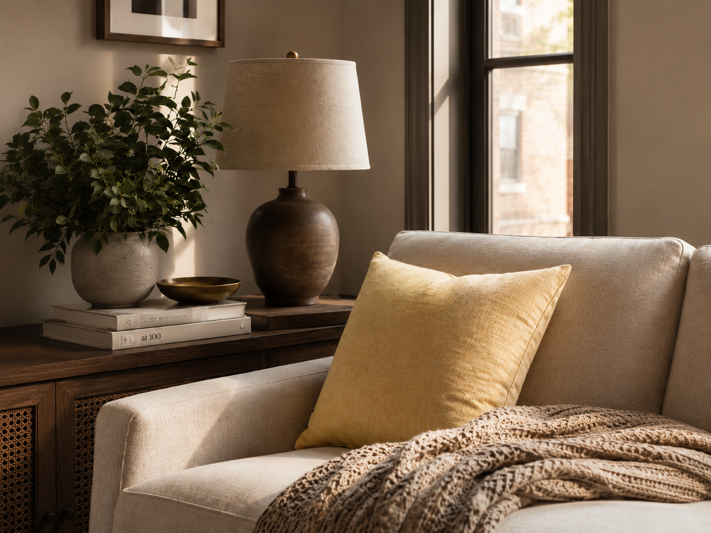 A cozy living room corner with a beige sofa, a yellow throw pillow, and a patterned blanket. Beside the sofa, there is a wooden side table with a potted plant, a ceramic table lamp, and a few stacked books. A window brings in natural light, illuminating the space.