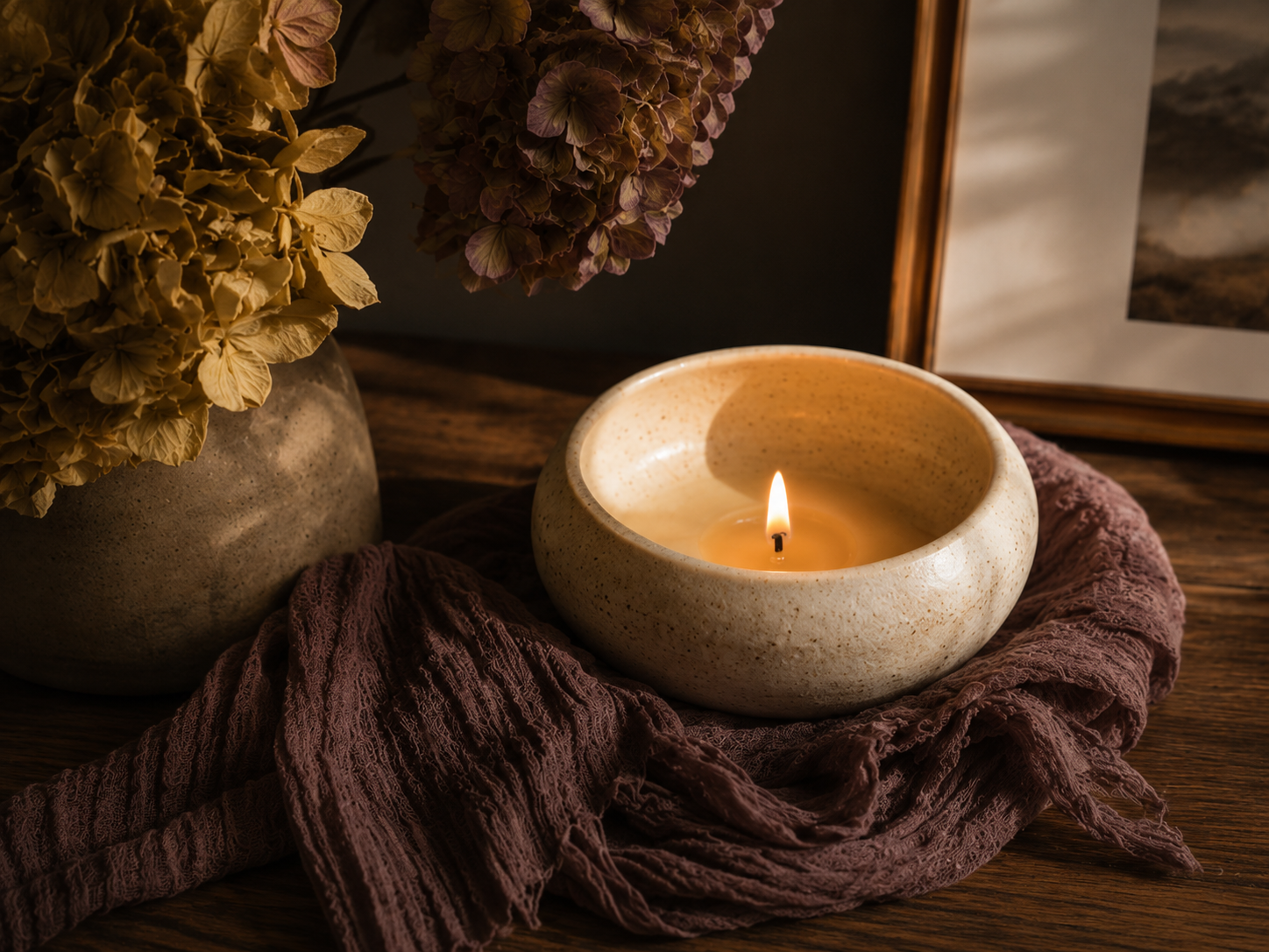 A lit candle in a cream-colored ceramic holder, surrounded by dried hydrangea flowers in a vase, with a framed picture and a mauve fabric on a wooden surface.