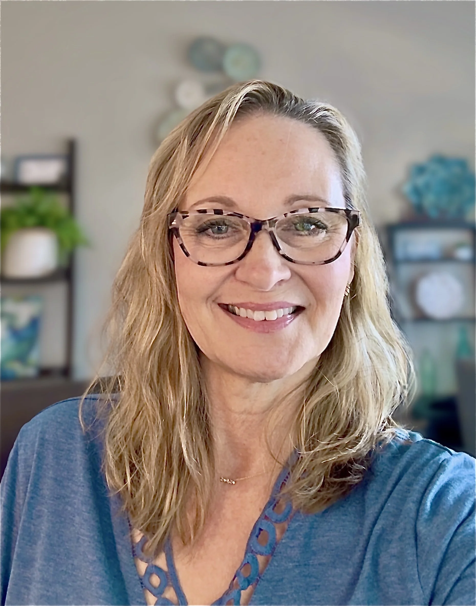 A woman with blonde, wavy hair and light skin wearing glasses and a blue top, smiling at the camera in a bright room with shelves and plants in the background.
