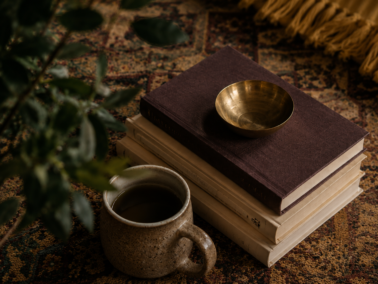 A stack of three books on a patterned rug, with a small brass bowl on top of the dark purple book, a ceramic mug filled with dark liquid in front of the books, and a green leafy plant partially visible on the left.