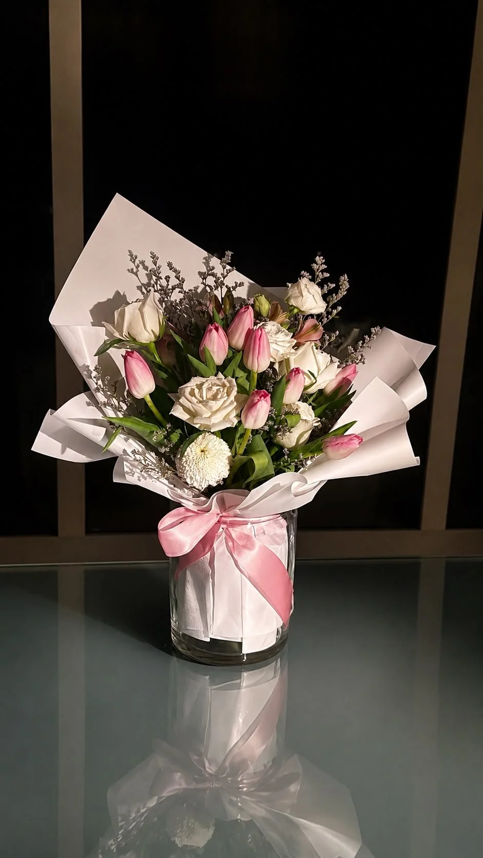 A bouquet of pink tulips, white roses, and white chrysanthemum in a glass vase with a pink ribbon, wrapped in white decorative paper, sitting on a glass table.