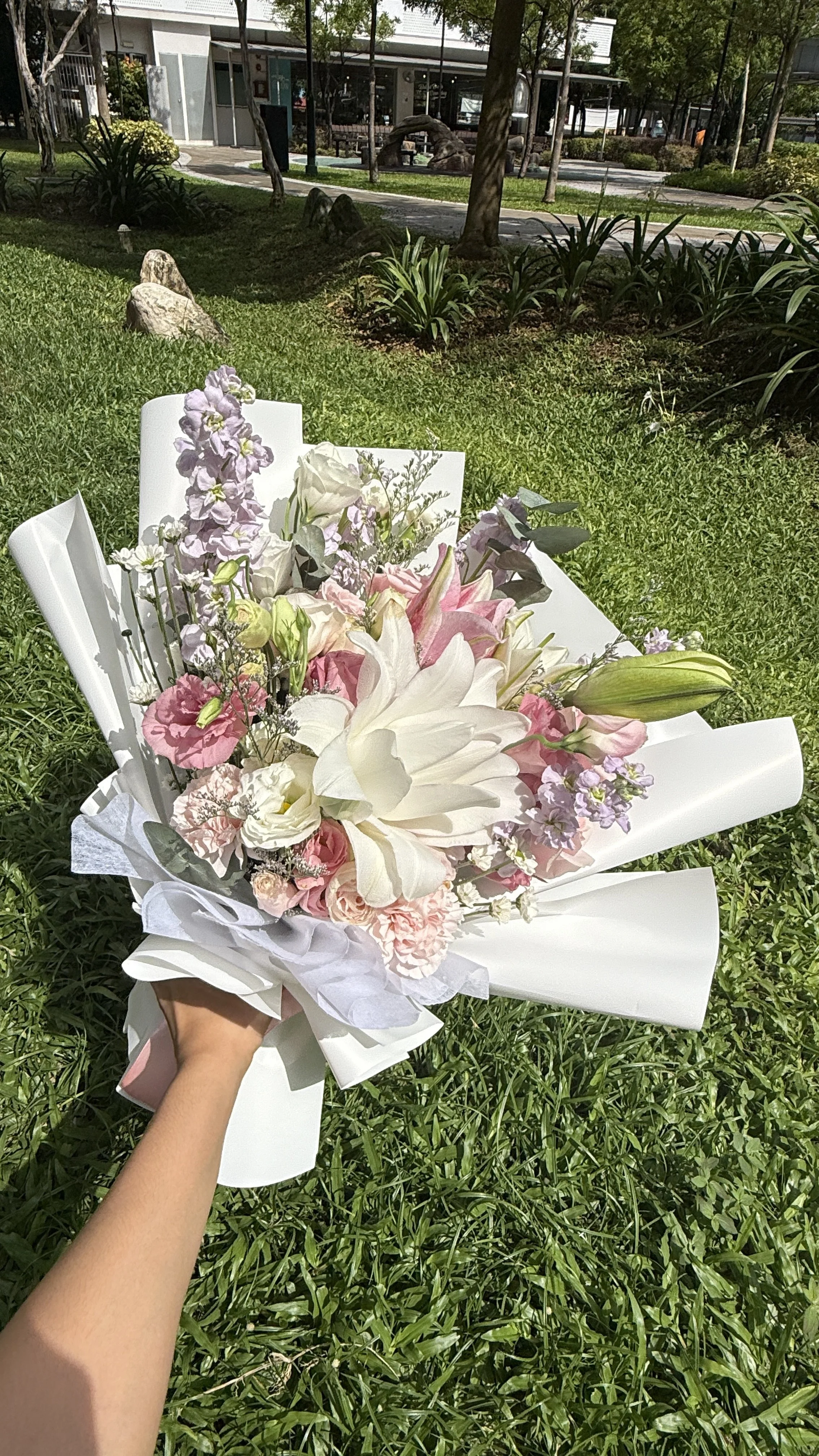 A colorful bouquet of flowers including white lilies, pink roses, lavender, and other blossoms being held outside in a park-like area with trees, grass, rocks, and a building in the background.