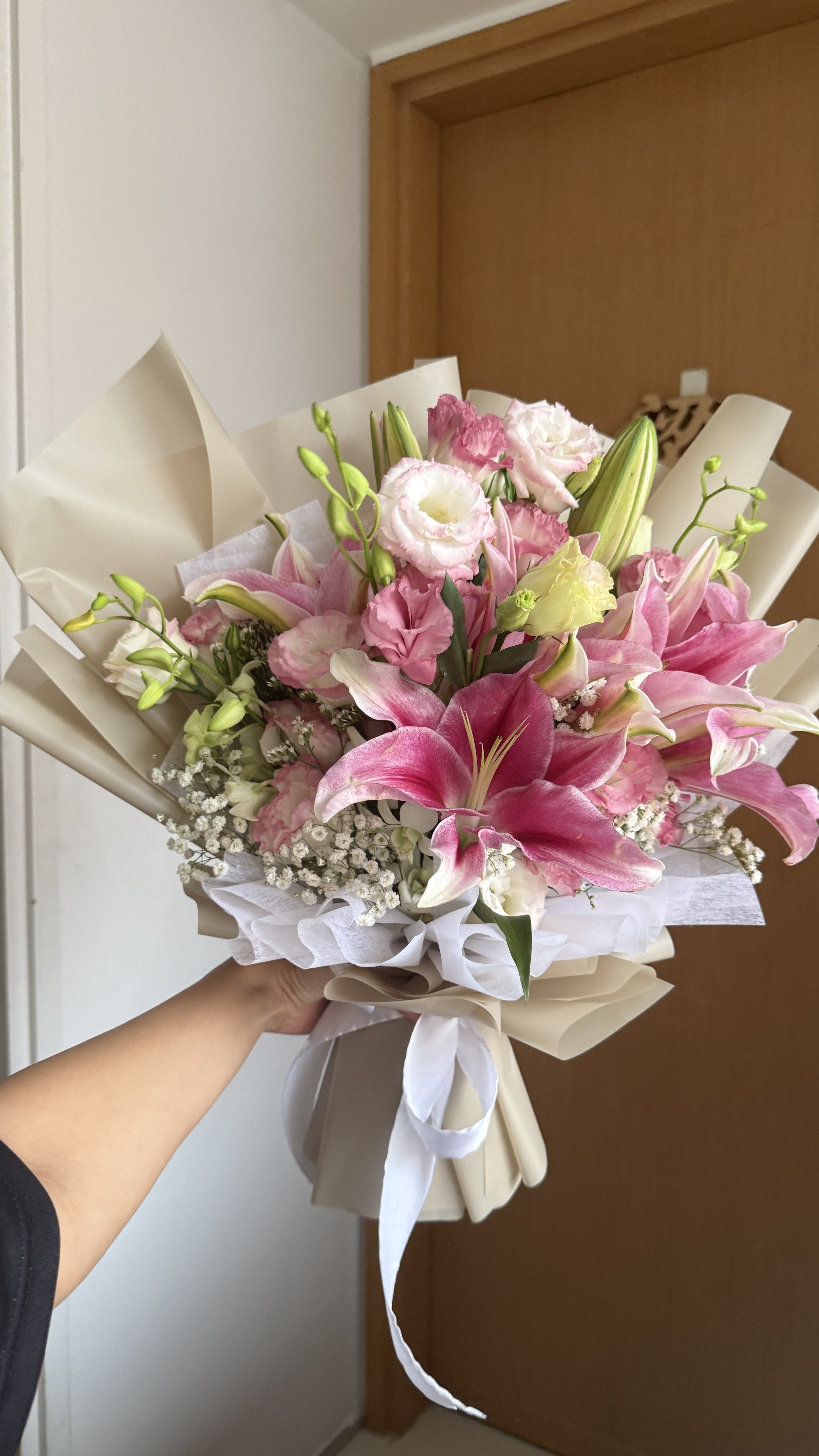 A bouquet of pink and white flowers, including lilies and other blooms, wrapped in white paper and tied with a white ribbon, held by a person's hand.
