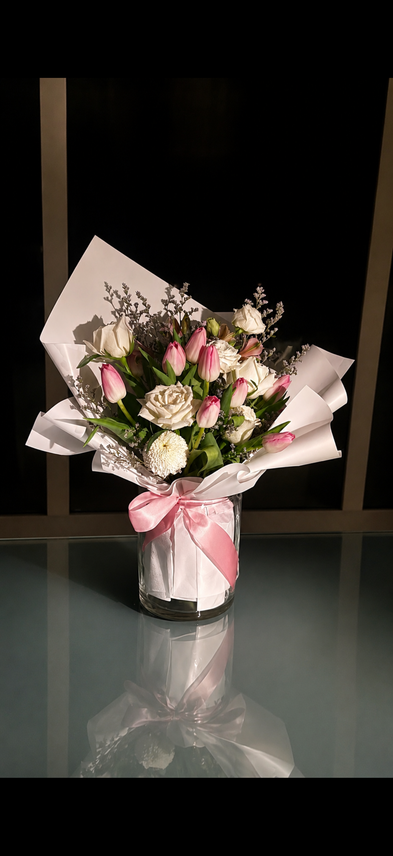 A bouquet of white and pink tulips, white roses, a white flower, and sprigs of lavender, wrapped in white paper with pink ribbon, in a glass vase on a glass table with a mirror reflection, against a dark background.
