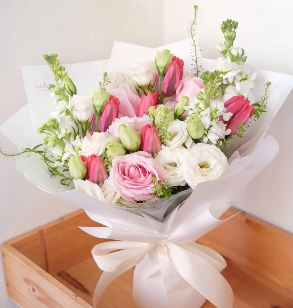 A bouquet of pink and white flowers wrapped in white paper and tied with a white ribbon, placed on a wooden surface against a light-colored wall.