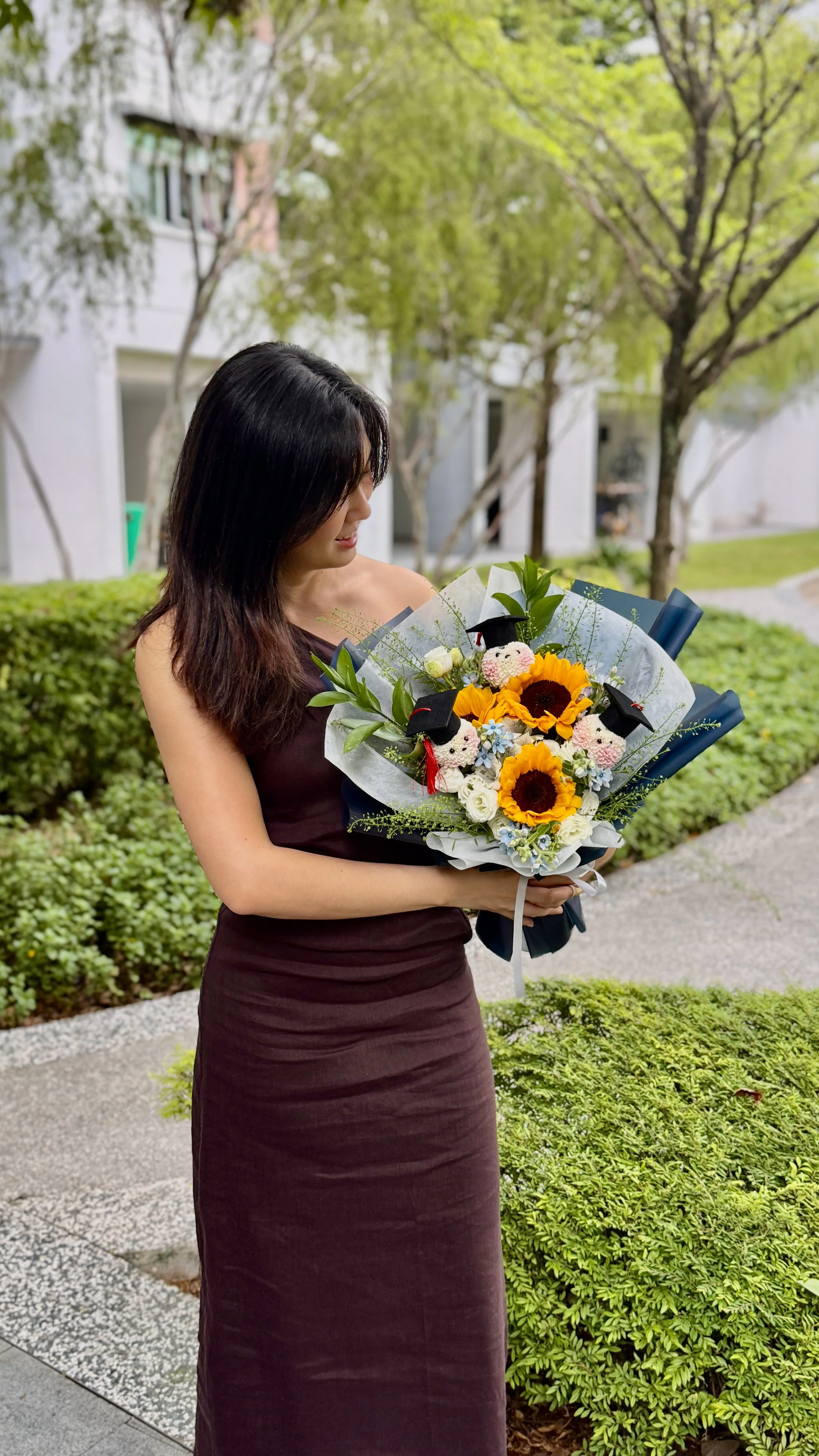 A woman in a sleeveless dark dress holding a bouquet of sunflowers, roses, and small stuffed sheep with graduation caps, outdoors with trees and bushes in the background.