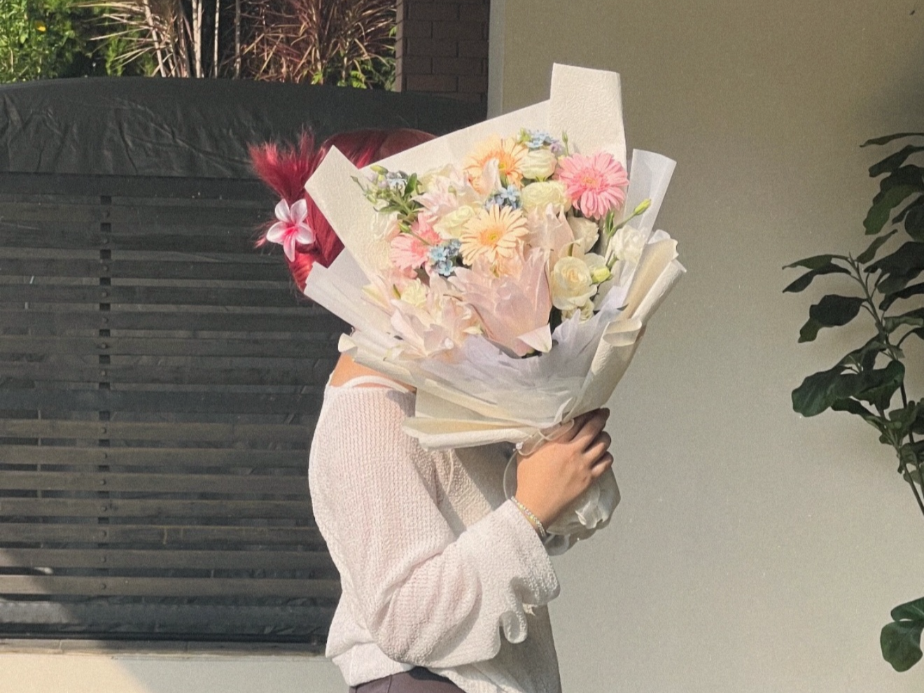 Person holding a large bouquet of mixed flowers covering their face, standing indoors near a wall and a green plant.