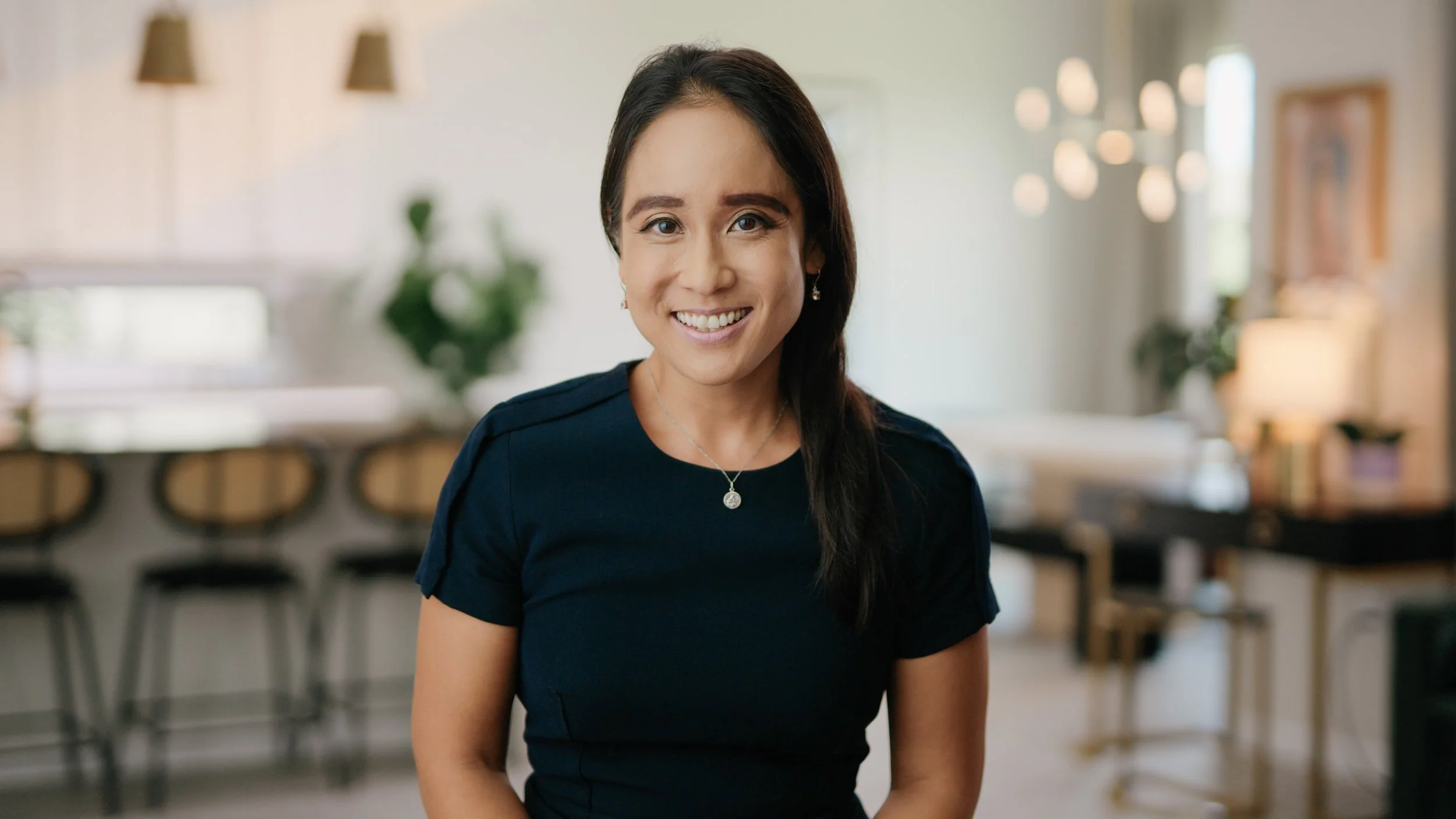 A smiling woman with long dark hair wearing a dark blue top and a silver necklace, standing in a bright, modern indoor space with blurred background.