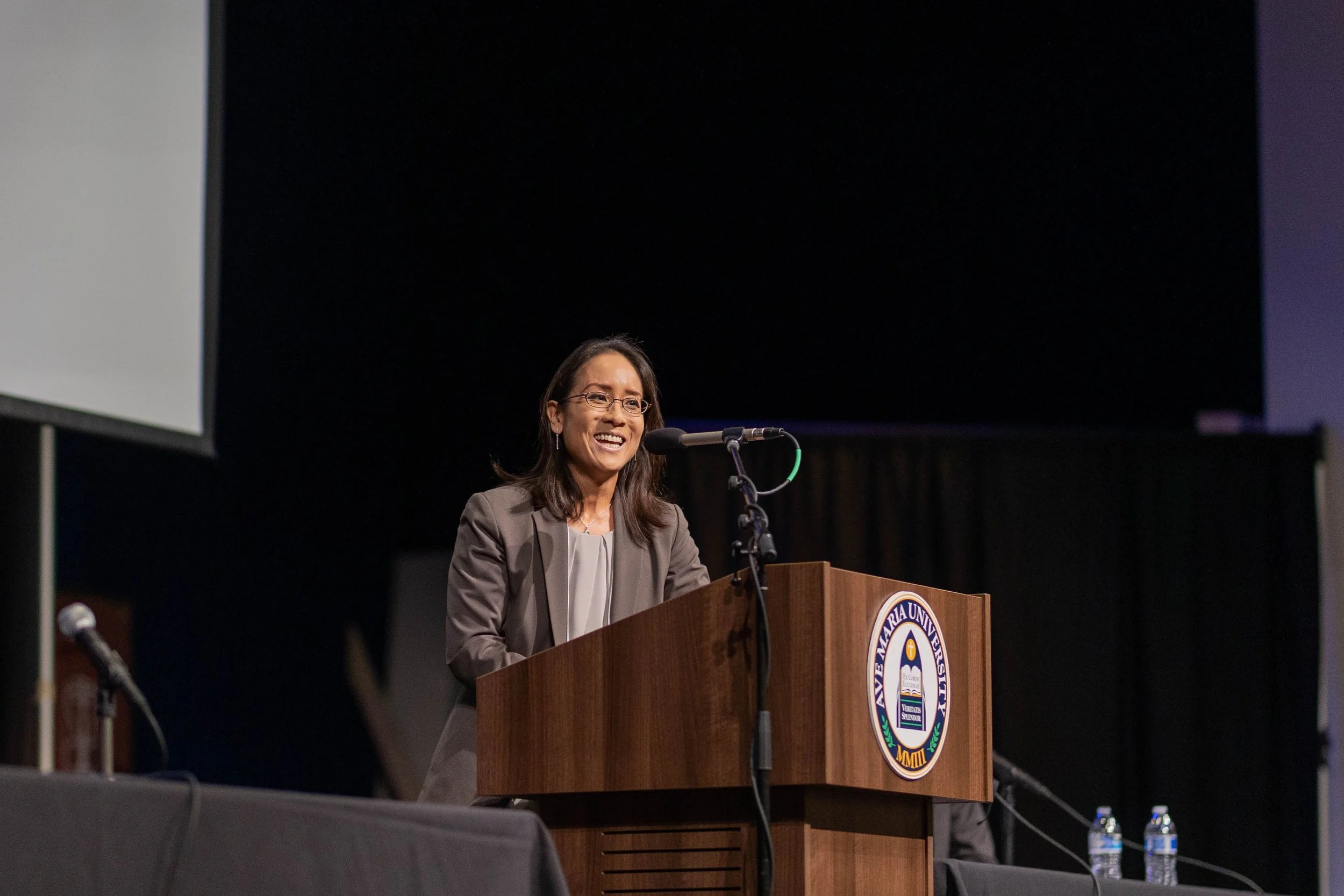 A woman standing behind a wooden podium with a seal, smiling and speaking at an event, with microphones and water bottles nearby.