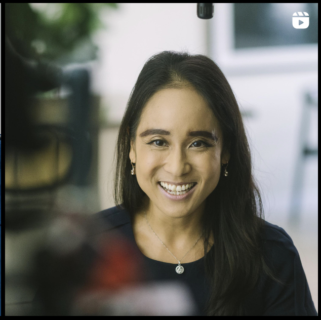 A woman with long dark hair smiling at the camera in a bright indoor setting.
