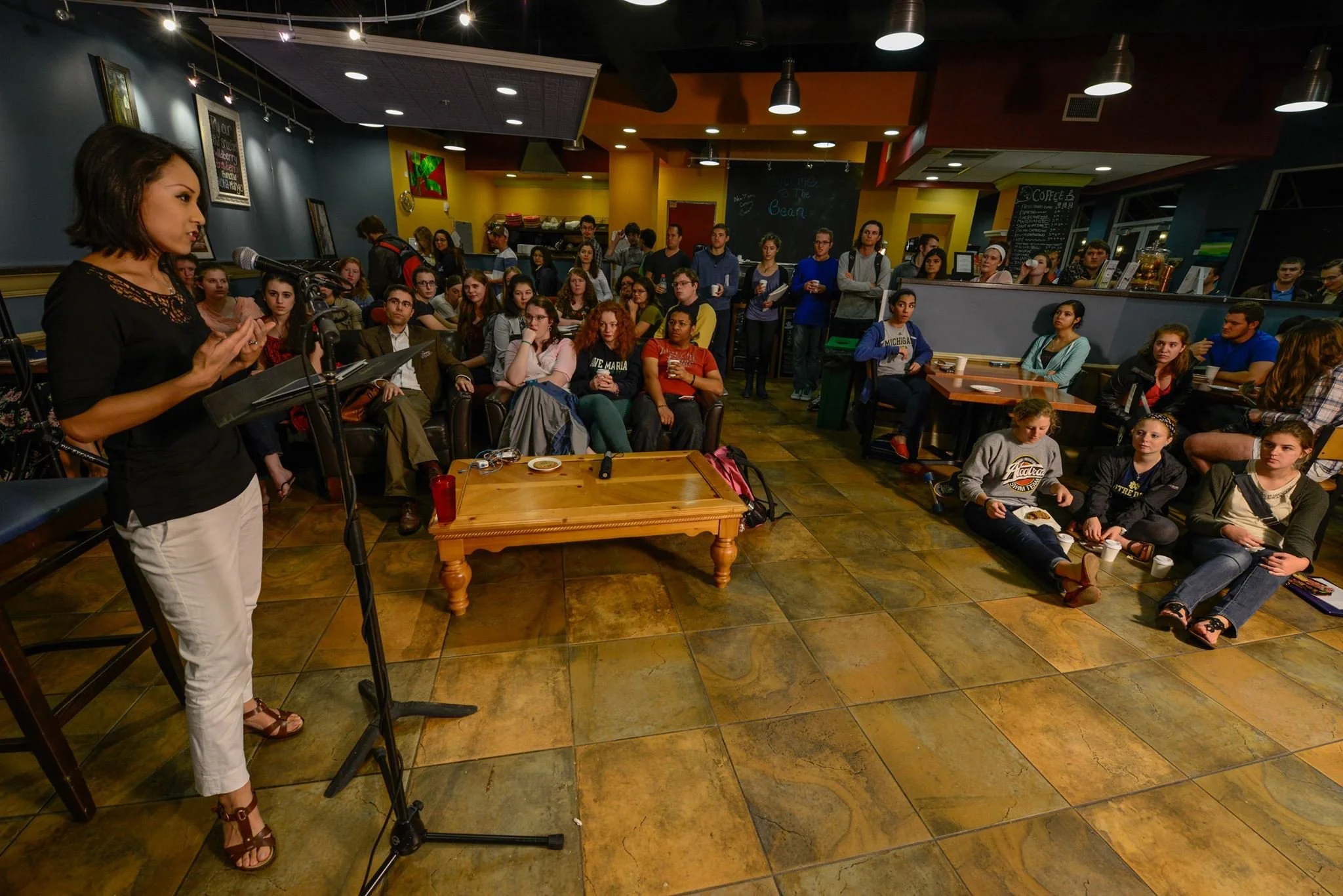 A woman with short dark hair, wearing a black top and white pants, is speaking into a microphone at a podium in a crowded cafe or coffee shop. The audience, consisting of young adults and some middle-aged individuals, is seated and standing, attentively listening. The background features colorful walls, artwork, a chalkboard menu, and coffee station.
