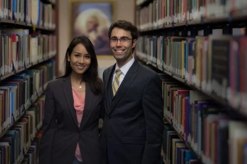 Two people, a woman and a man, smiling in a library aisle with bookshelves on both sides.