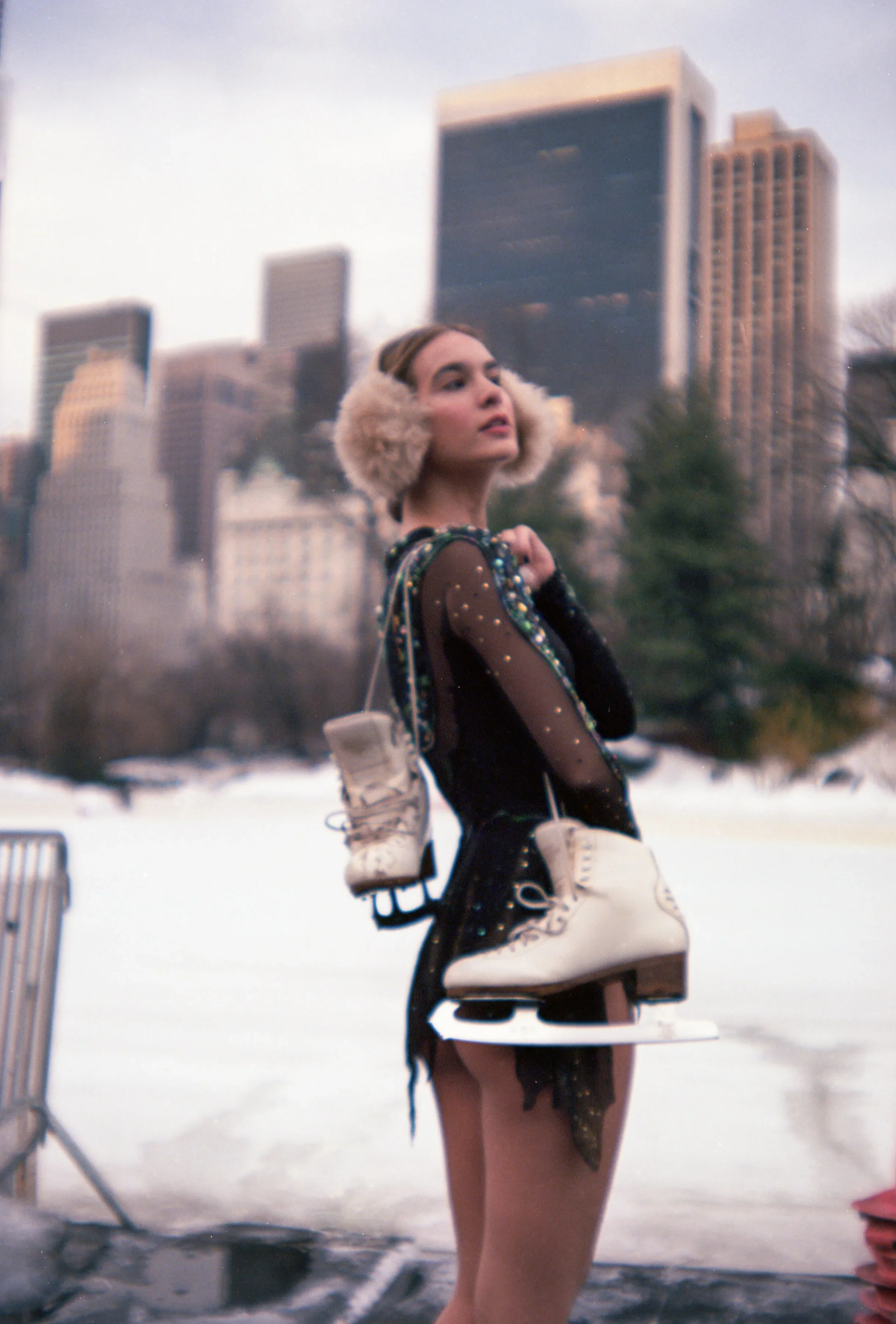 Female figure skater wearing an embellished skating dress and earmuffs, holding white ice skates in Central Park, NYC with Manhattan skyline background, cinematic winter photography by Lenny Kats