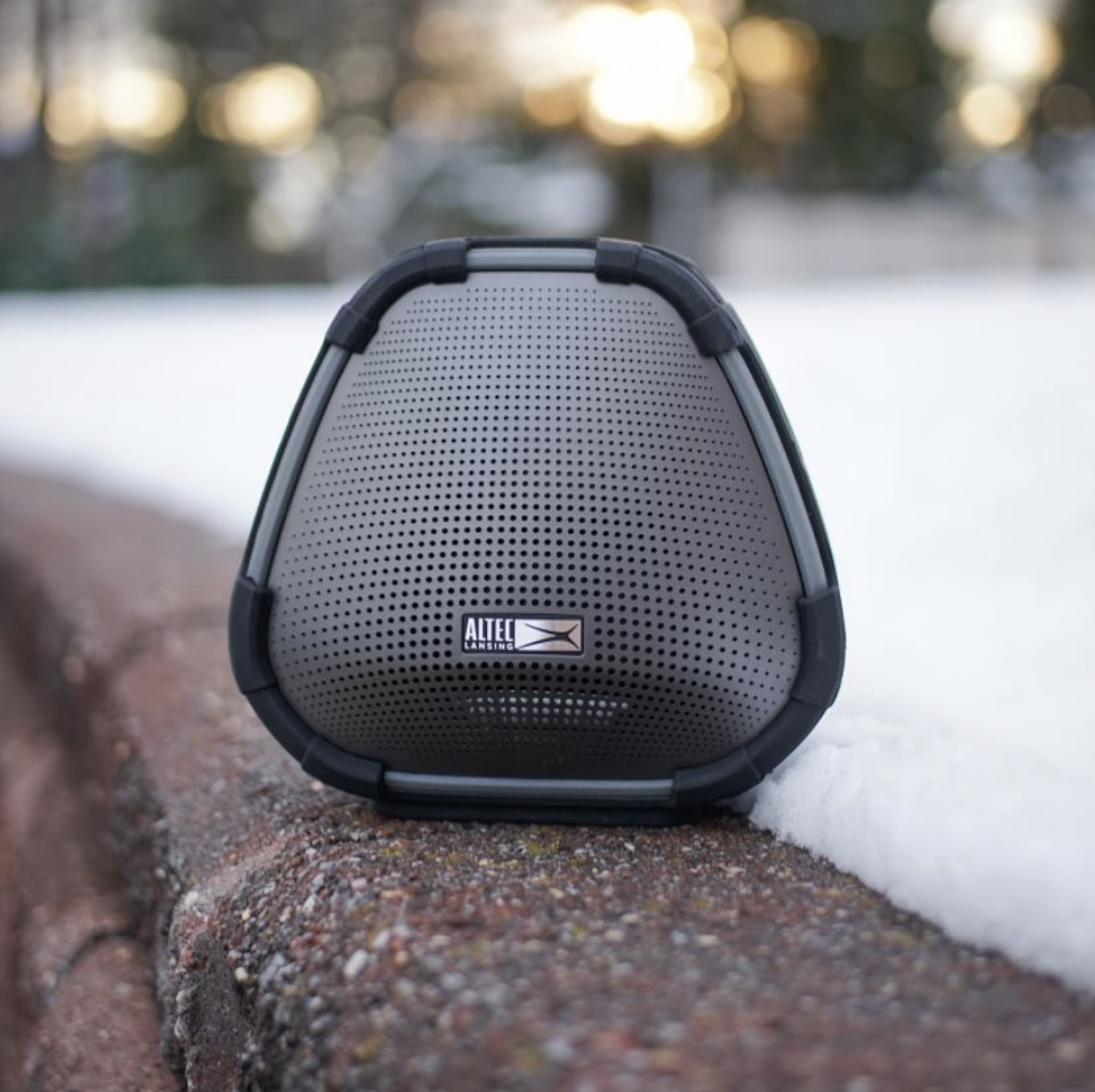 Outdoor scene with a portable speaker placed on a brick ledge, snow on the ground, and blurred trees and sunset in the background.