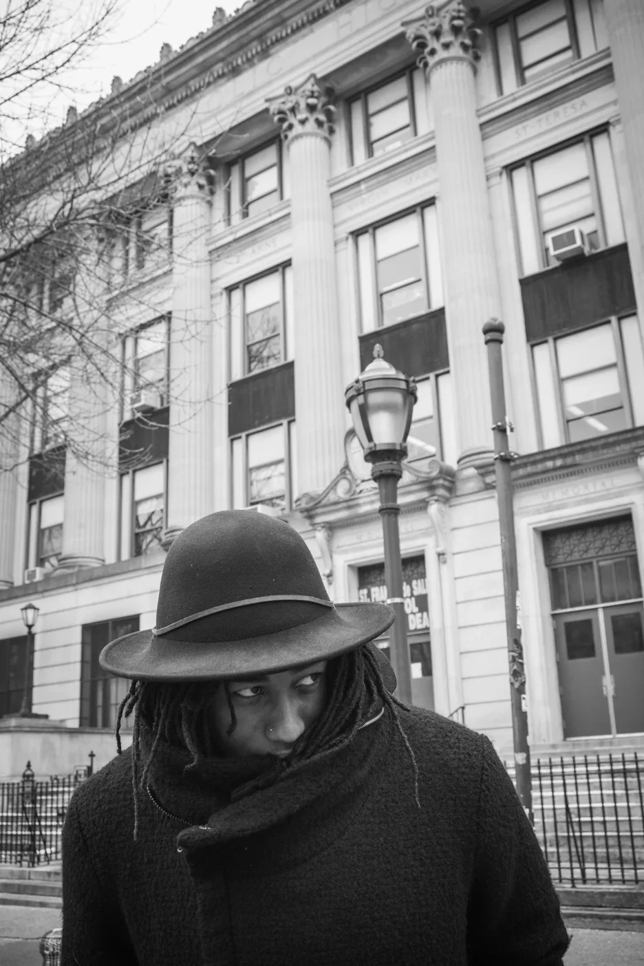 Black and white editorial street portrait of a model with dreadlocks wearing a fedora hat and scarf in Brooklyn, NYC. Photographed by Leonard 'Lenny Kats' Katsel against a historic building with classical columns and urban architecture.