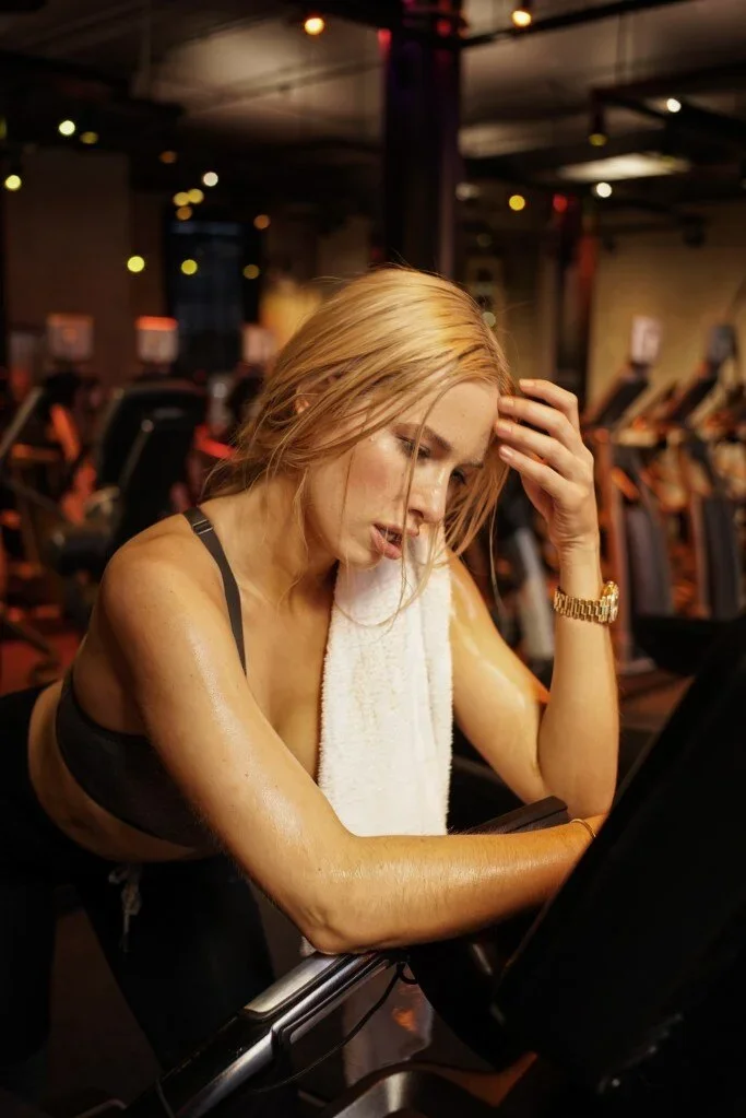 Doroshina (@doroshina) blonde model sweating after intense gym workout at NYSC for Come Dry Leave Wet campaign, leaning on cycling bike with towel and gold watch, NYC fitness photography