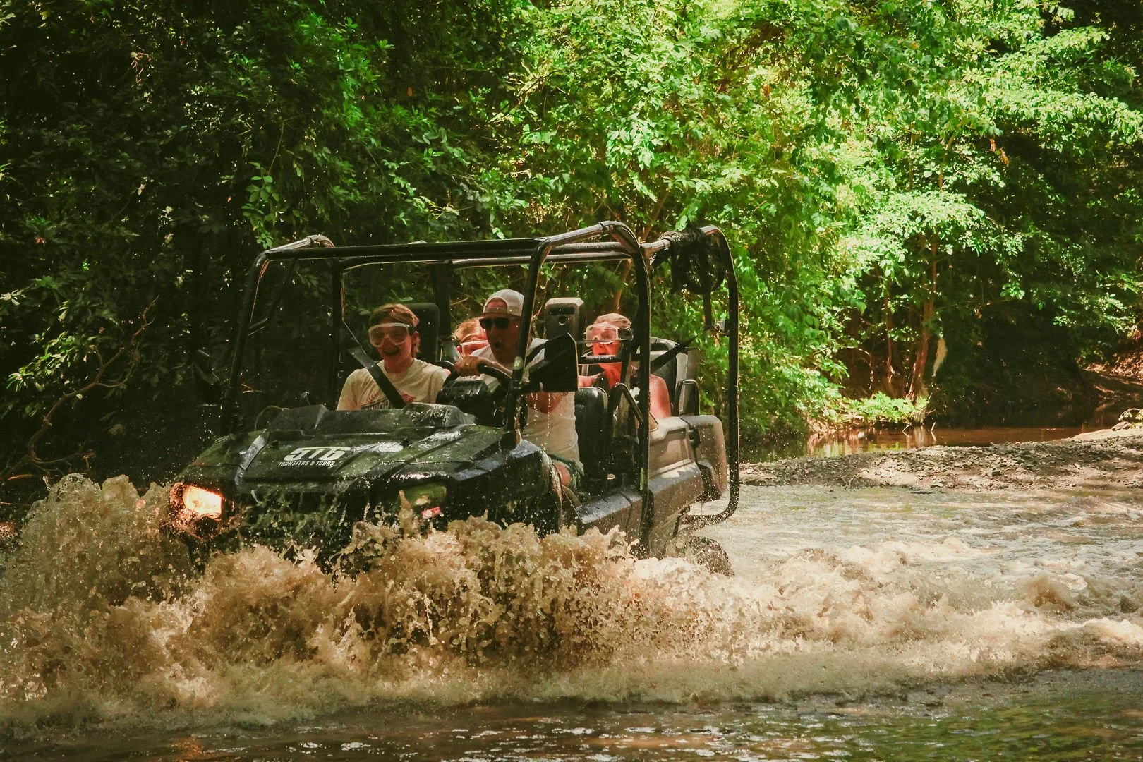 People riding an ATV through a shallow, muddy stream surrounded by lush green trees.