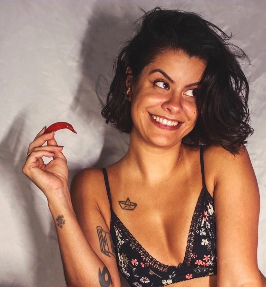 A young woman with short, dark, curly hair smiling and holding a red chili pepper. She is wearing a black floral lace camisole and has tattoos on her arm and chest.