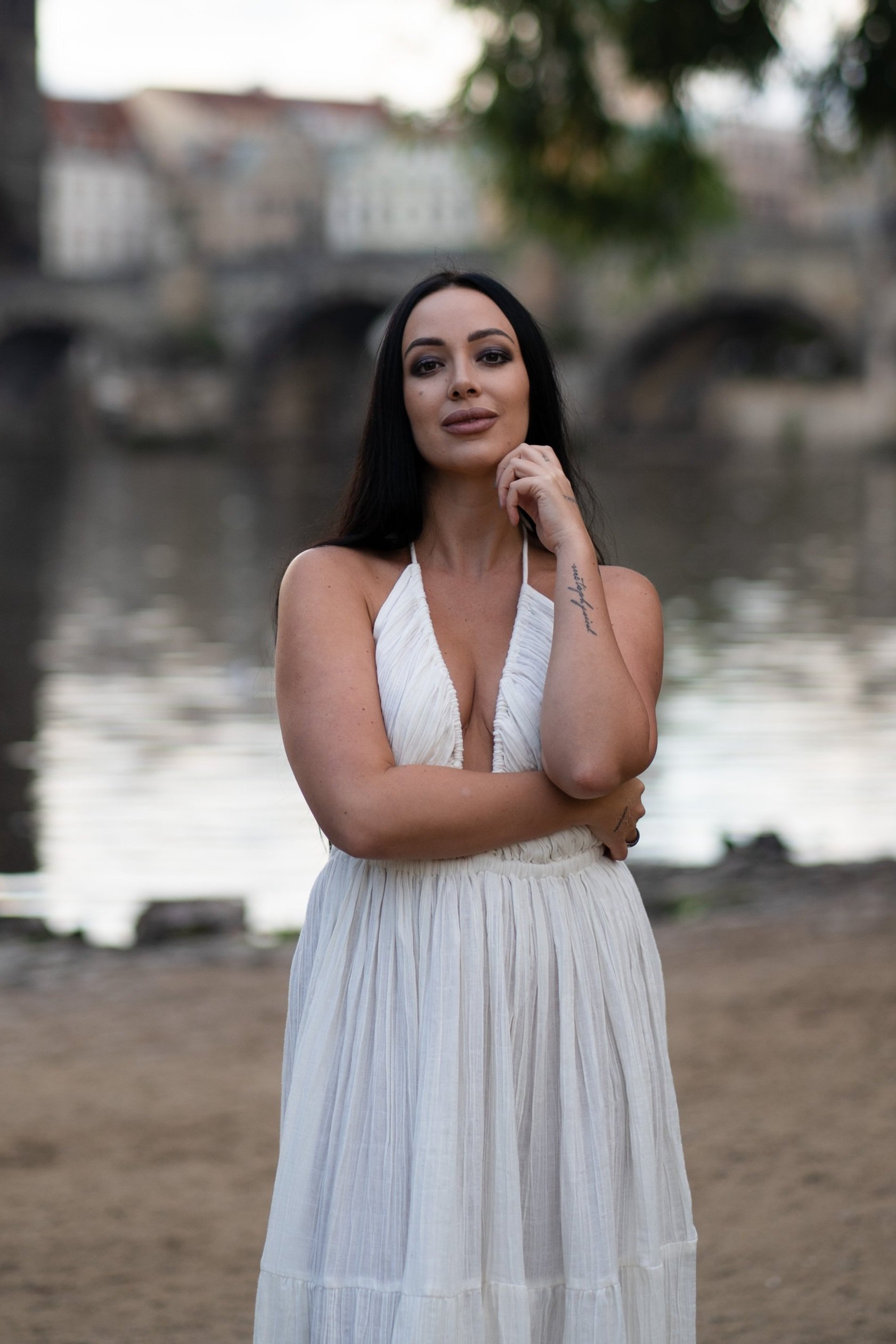 A woman with long black hair wearing a white dress standing outdoors near a river with a historic stone bridge in the background.