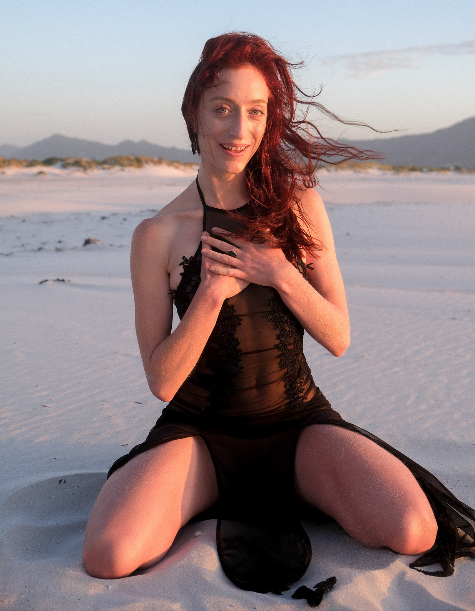 A woman with red hair kneeling on a sandy beach, wearing a black sheer dress, with mountains in the background during sunset.