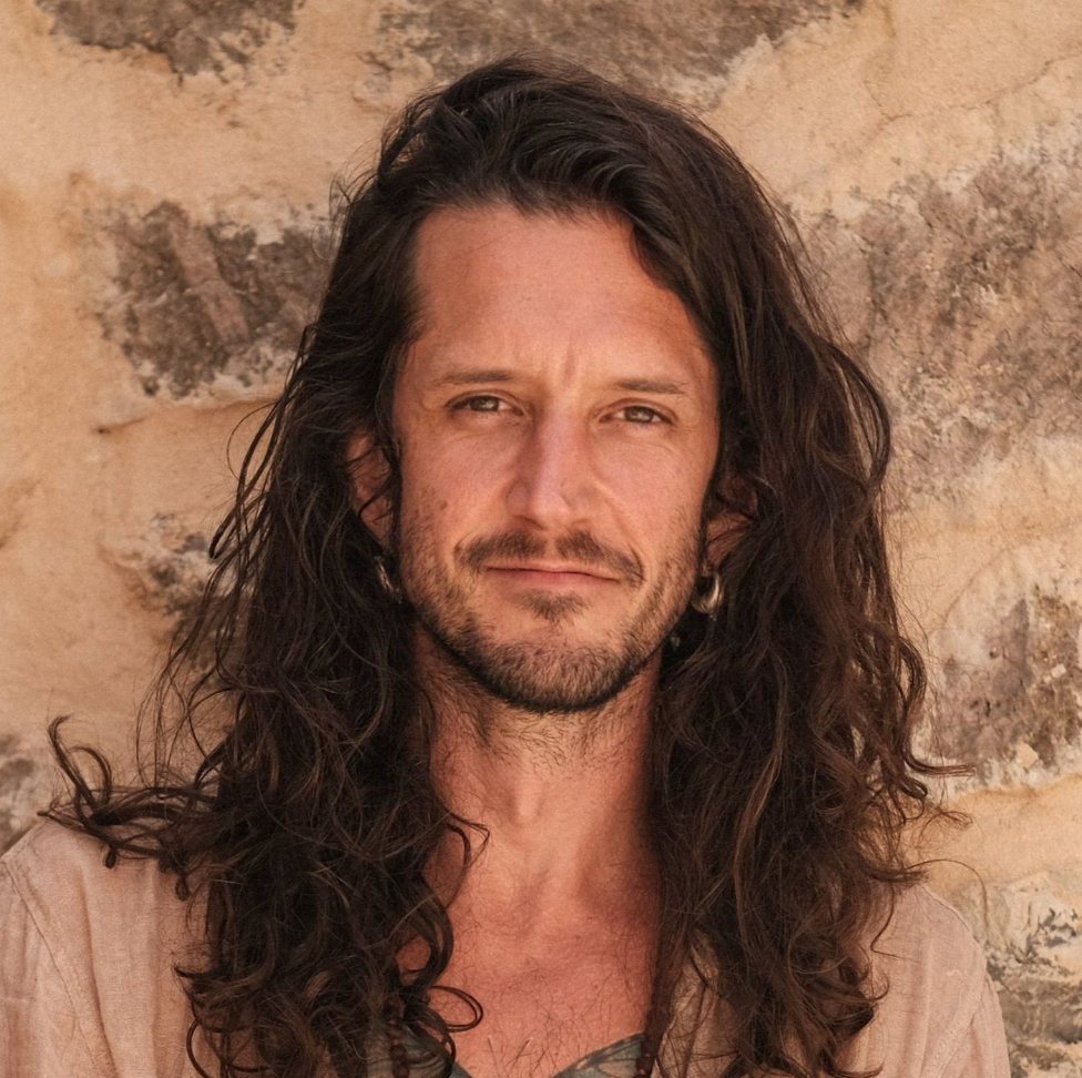 A man with long, wavy brown hair and a beard, standing against a textured stone wall