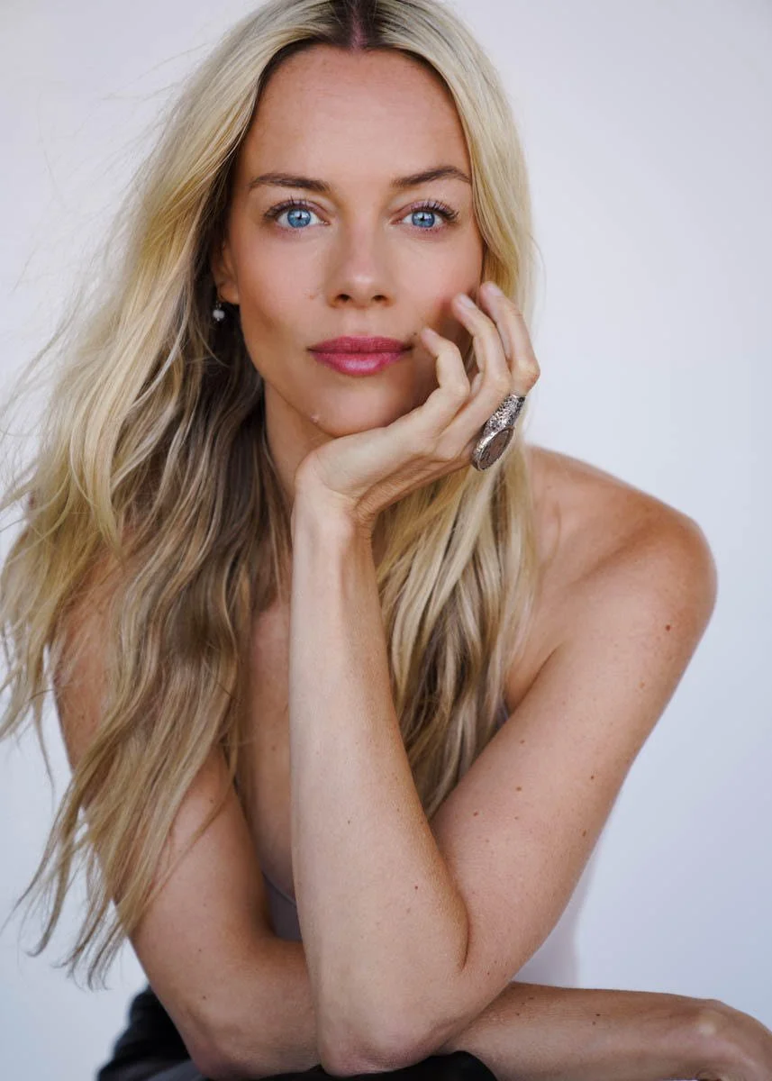 A woman with long blonde hair and blue eyes resting her face on her hand, looking at the camera with a slight smile, wearing a ring and earrings.